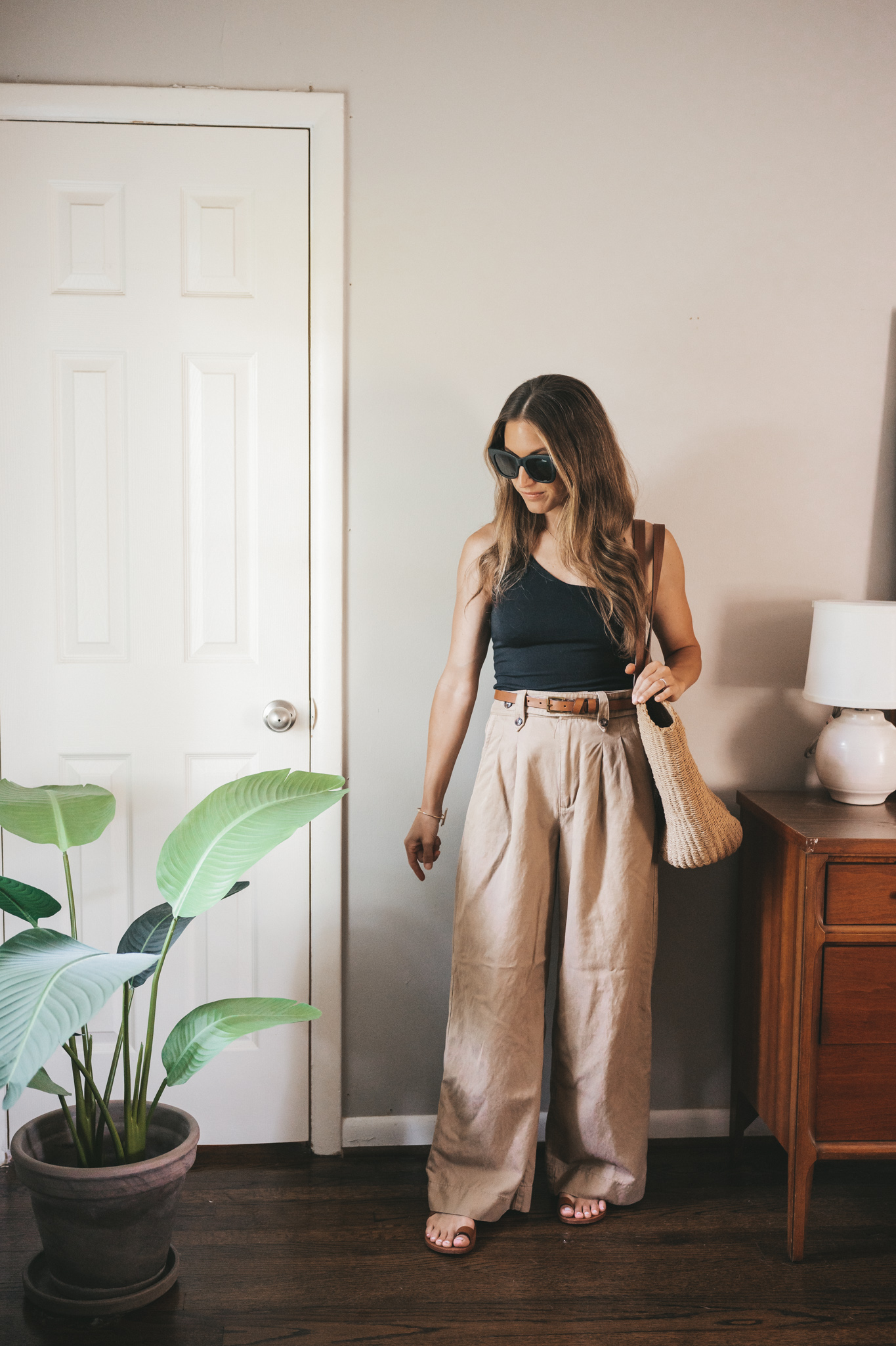 ashley wearing a black one shoulder tank, tan wide leg trousers, brown leather toe ring sandals, and black square frame sunglasses