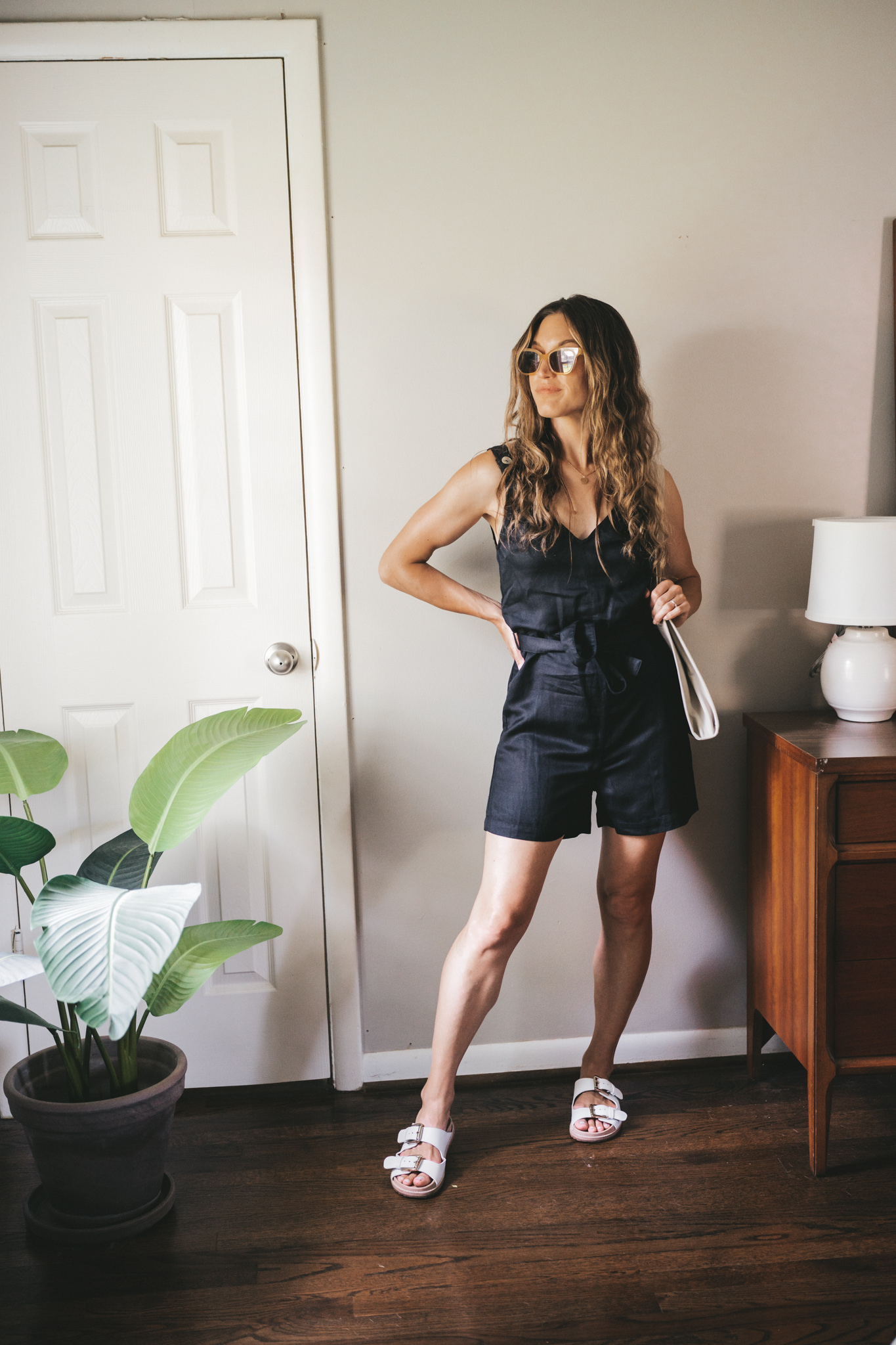 ashley wearing a black linen romper tied at the waist, white sandals, a gray shoulder bag, and tan frame sunglasses