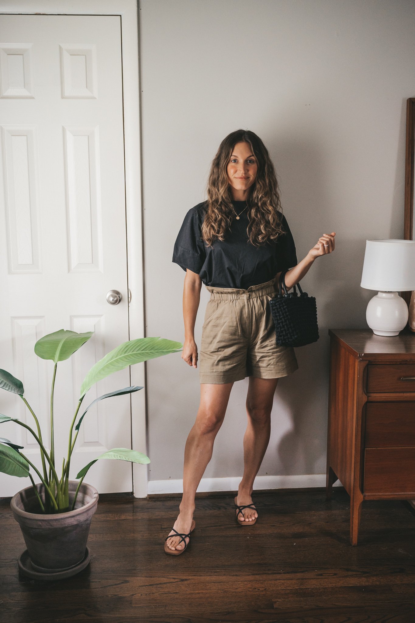 ashley wearing a black blouse, olive green paperbag shorts, black strappy sandals, a gold pendant necklace, and a black woven bag