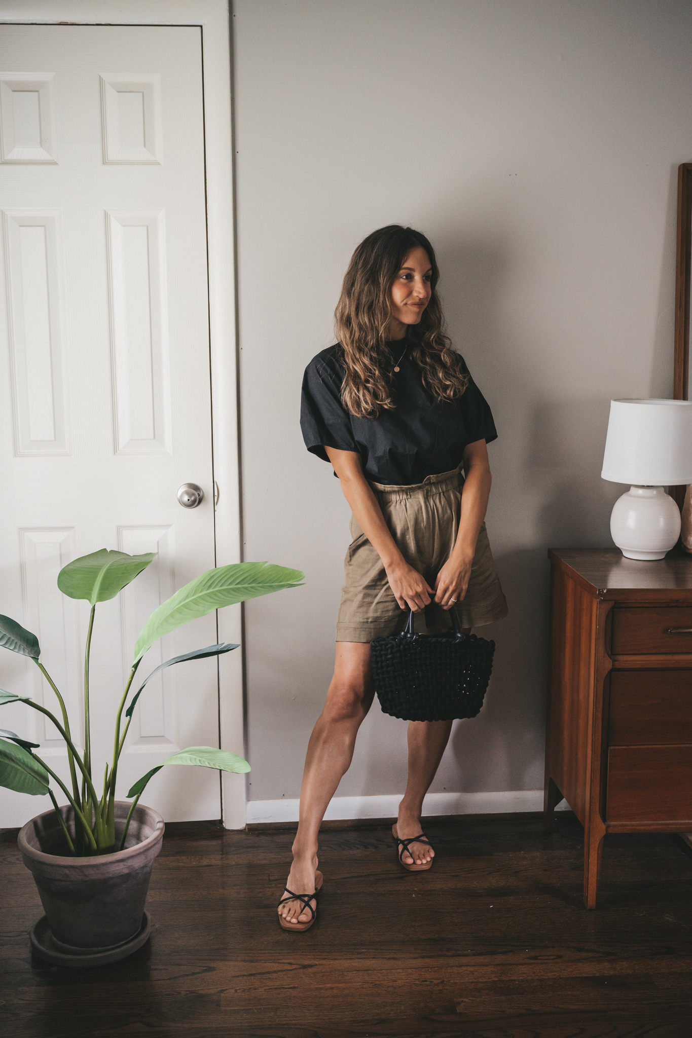ashley wearing a black blouse, olive green paperbag shorts, black strappy sandals, a gold pendant necklace, and a black woven bag