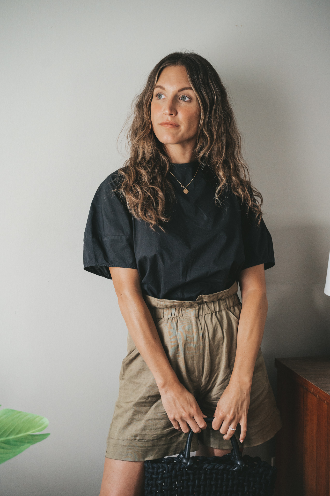 ashley wearing a black blouse, olive green paperbag shorts, black strappy sandals, a gold pendant necklace, and a black woven bag
