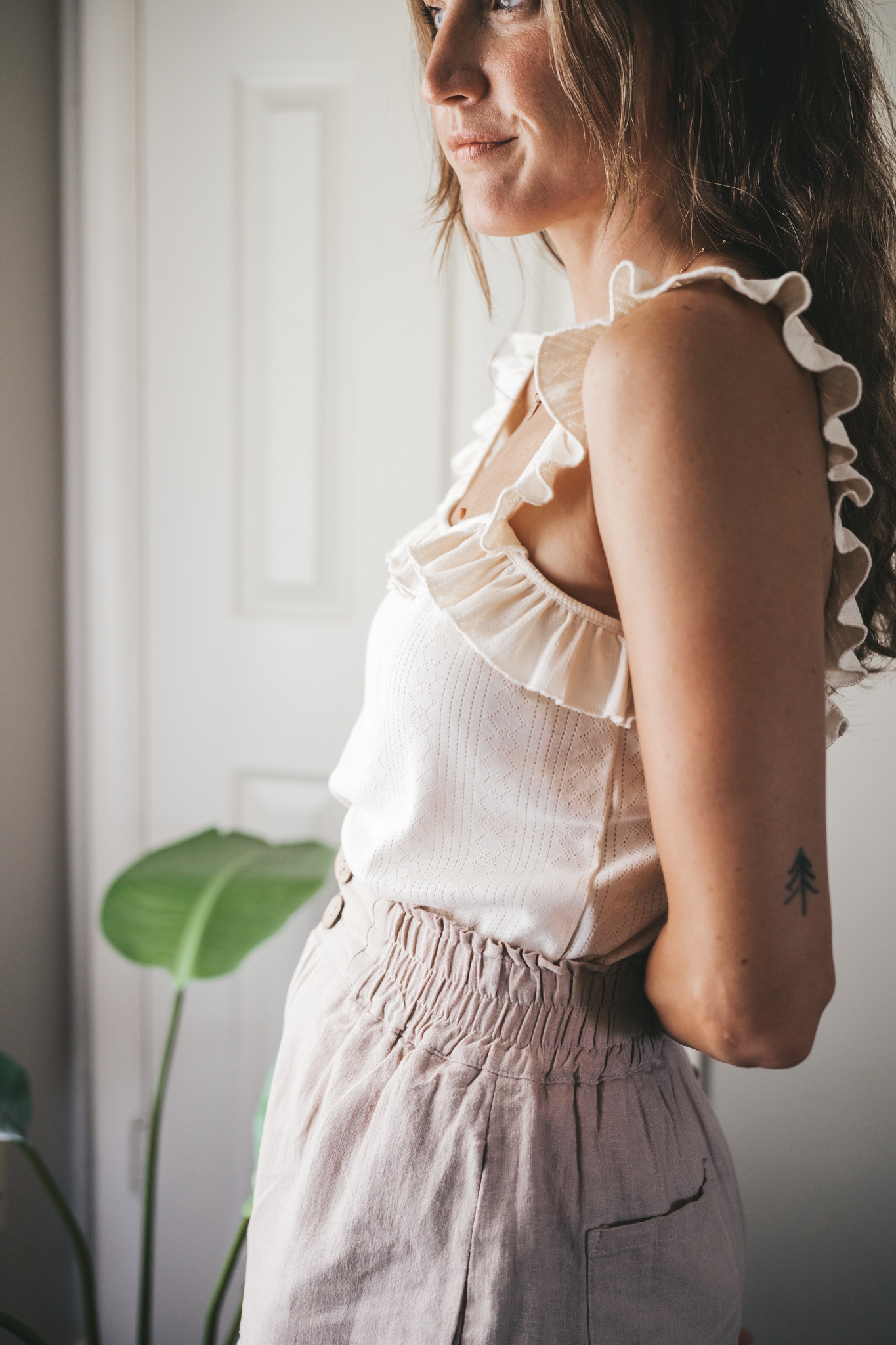ashley wearing a cream tank with ruffles on the straps, tan linen shorts, nude sandals, a woven bag, and tortoise shell sunglasses