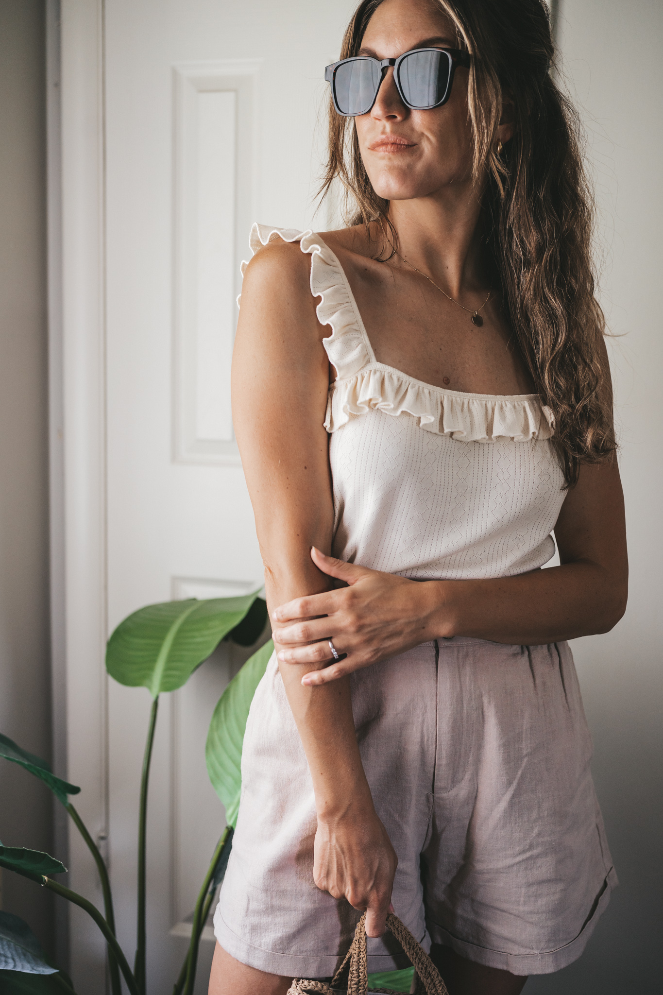 ashley wearing a cream tank with ruffles on the straps, tan linen shorts, nude sandals, a woven bag, and tortoise shell sunglasses