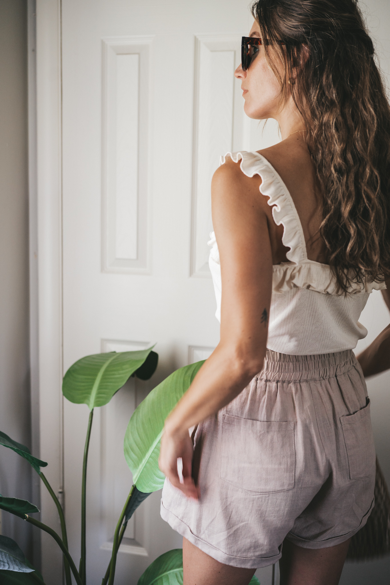 ashley wearing a cream tank with ruffles on the straps, tan linen shorts, nude sandals, a woven bag, and tortoise shell sunglasses