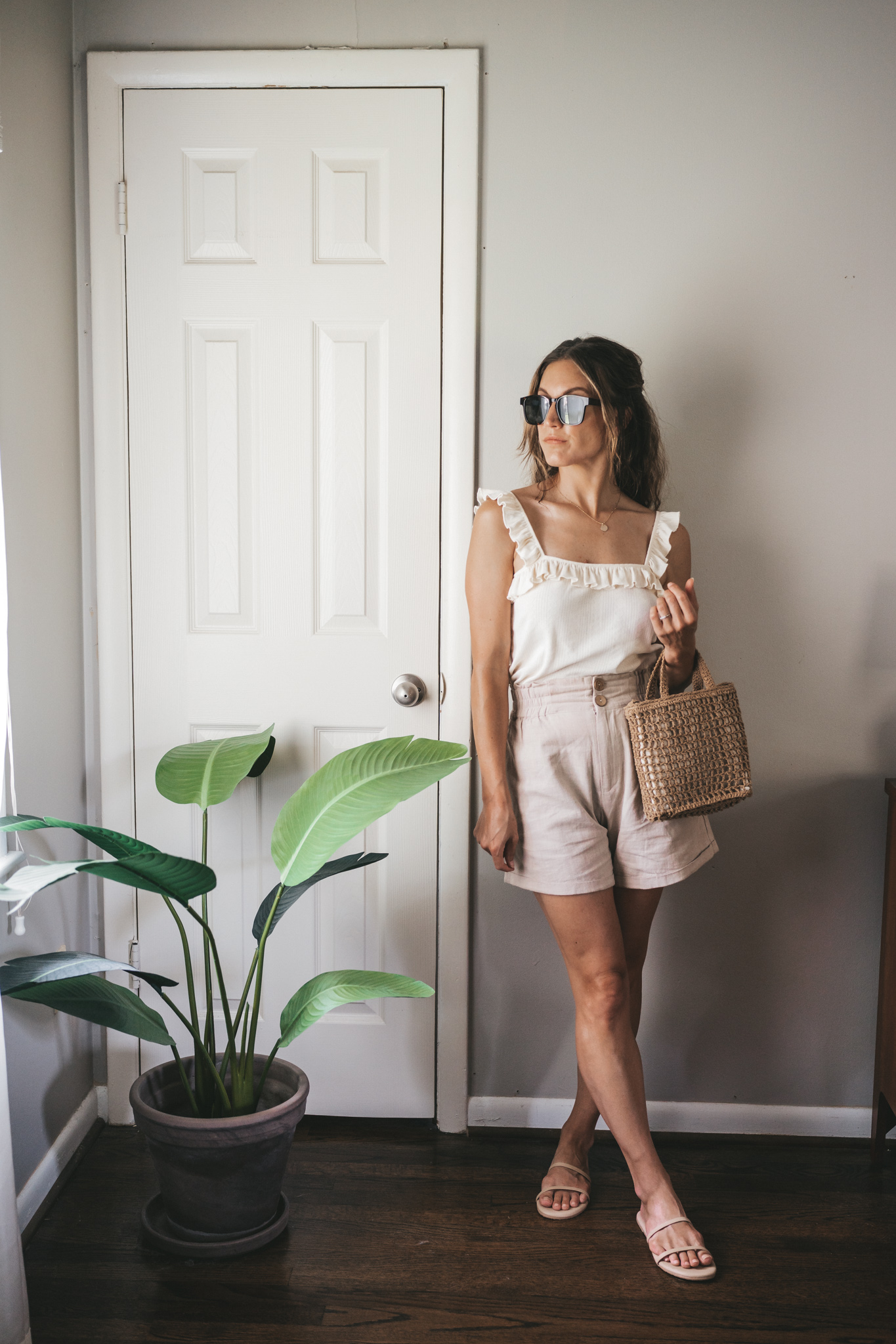 ashley wearing a cream tank with ruffles on the straps, tan linen shorts, nude sandals, a woven bag, and tortoise shell sunglasses