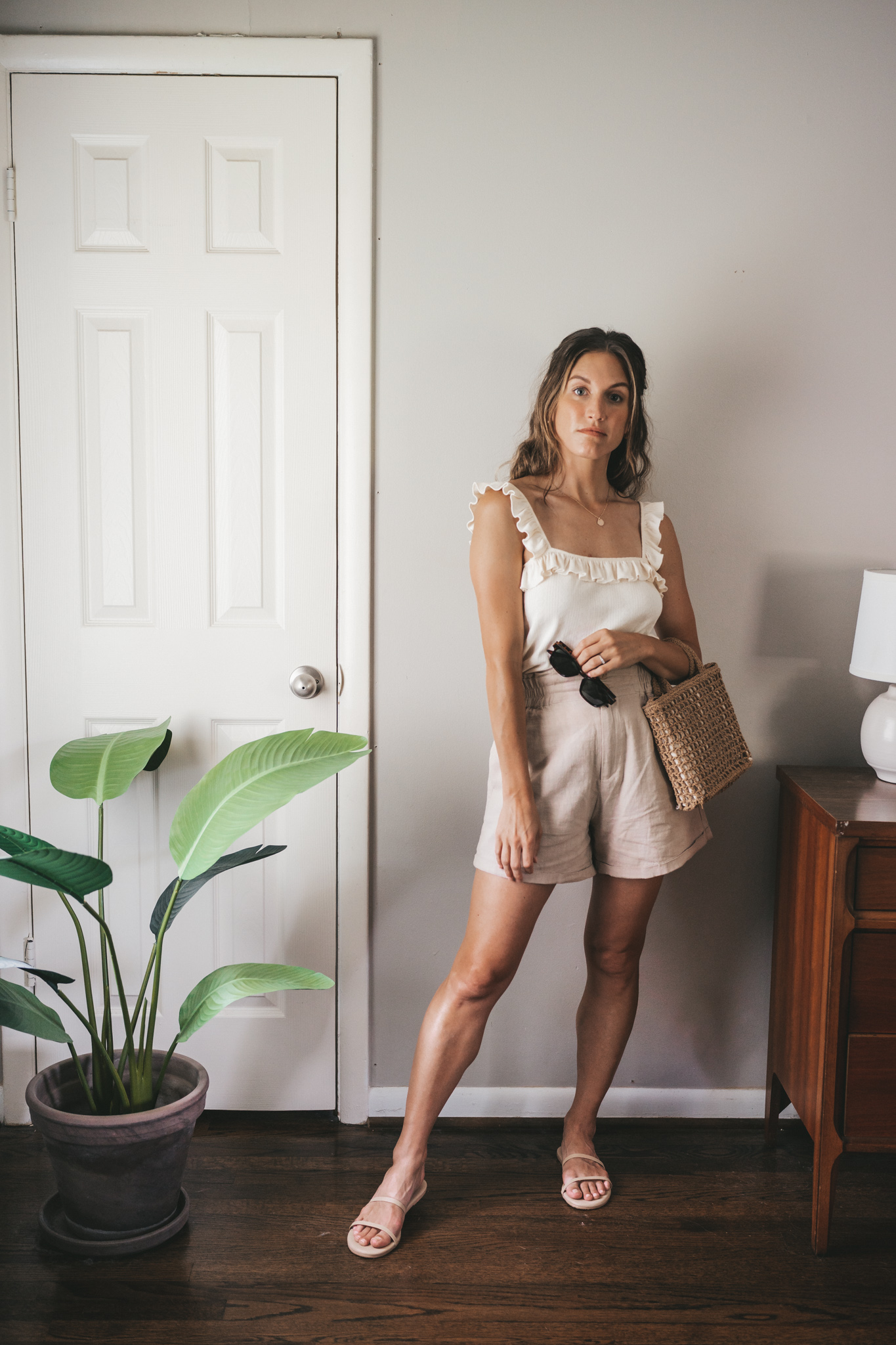 ashley wearing a cream tank with ruffles on the straps, tan linen shorts, nude sandals, a woven bag, and tortoise shell sunglasses