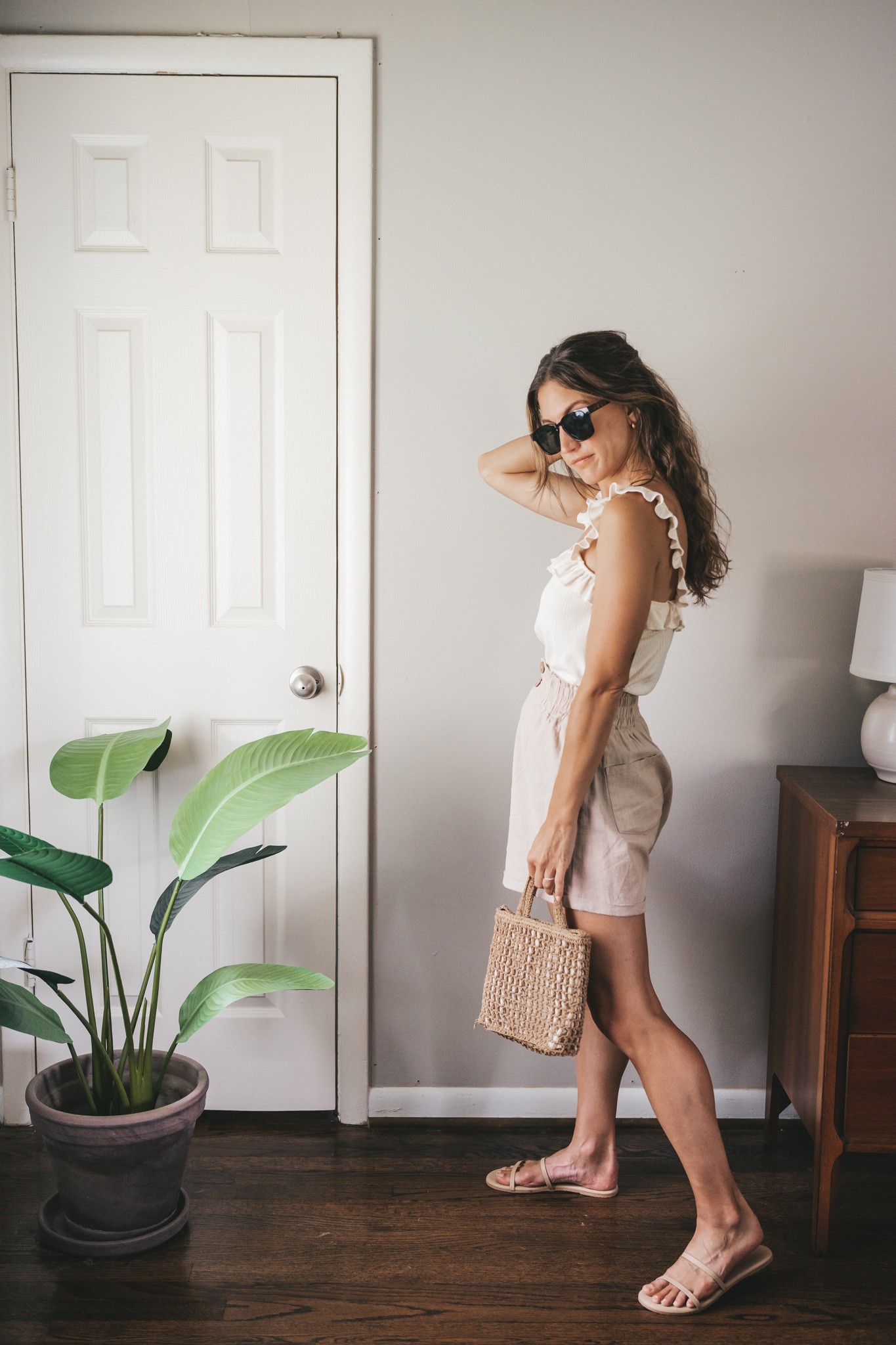 ashley wearing a cream tank with ruffles on the straps, tan linen shorts, nude sandals, a woven bag, and tortoise shell sunglasses