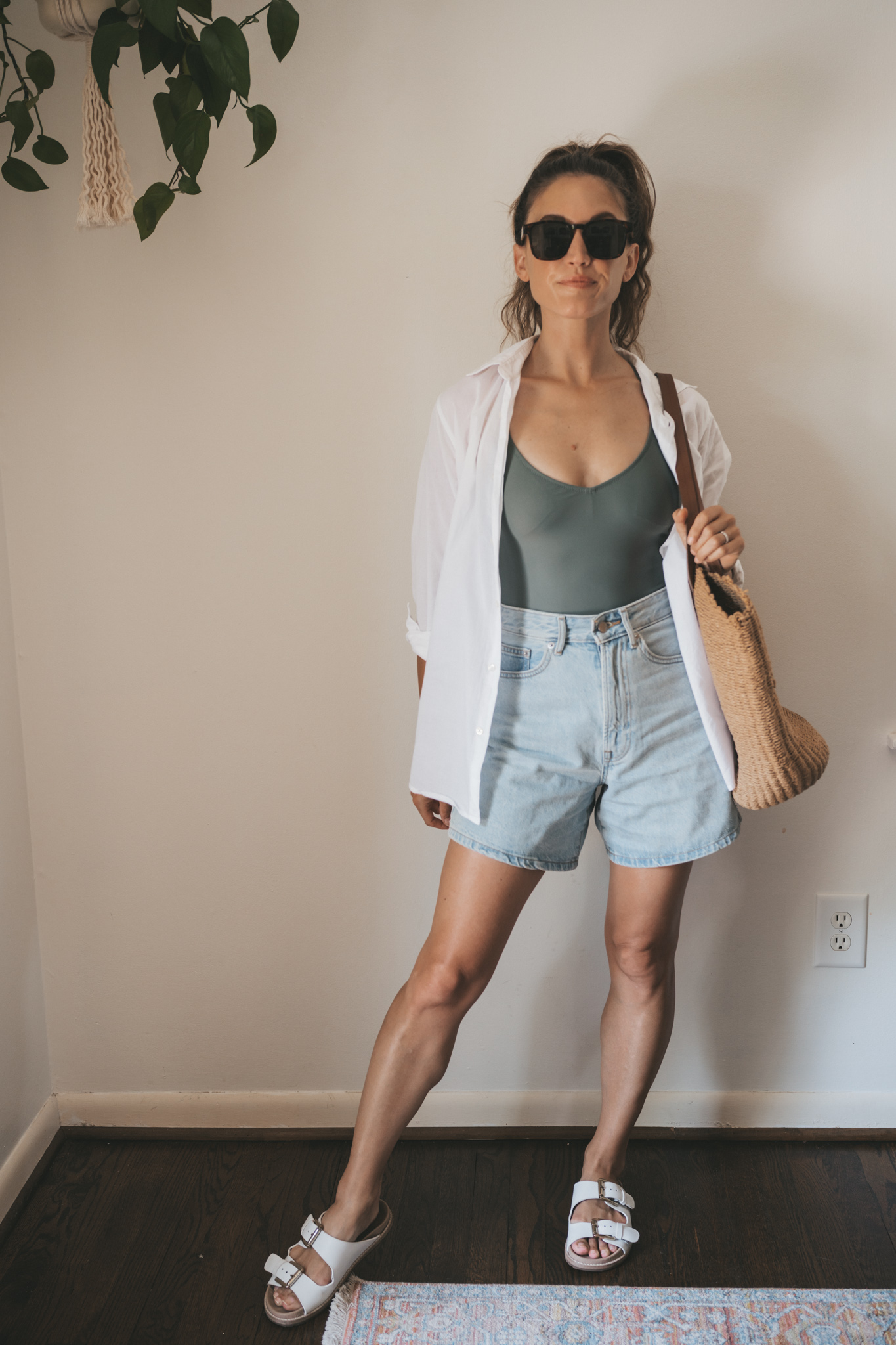 ashley wearing an olive green one piece swimsuit under a white button down, light wash denim shorts, and white sandals with tortoise shell sunglasses