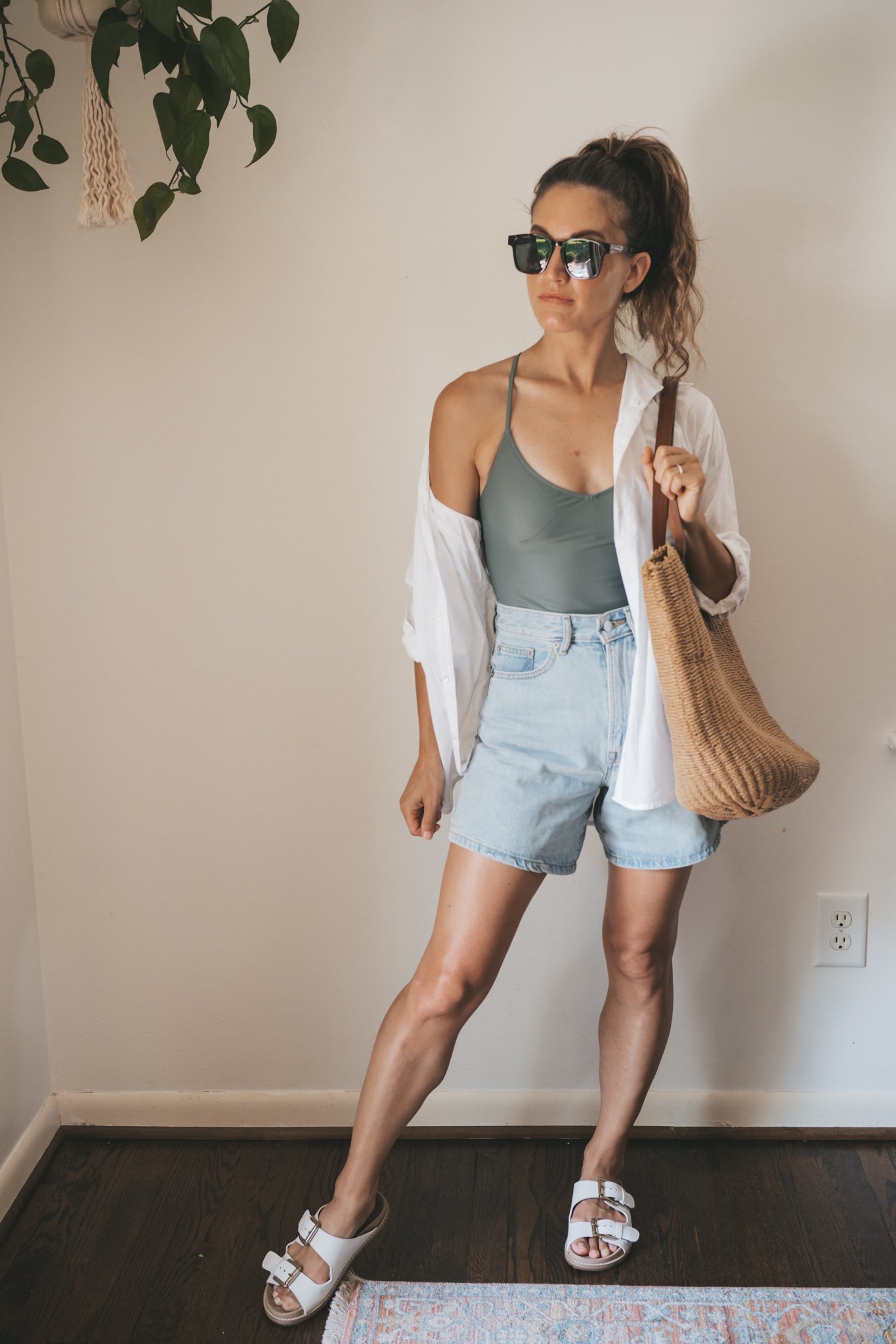 ashley wearing an olive green one piece swimsuit under a white button down, light wash denim shorts, and white sandals with tortoise shell sunglasses