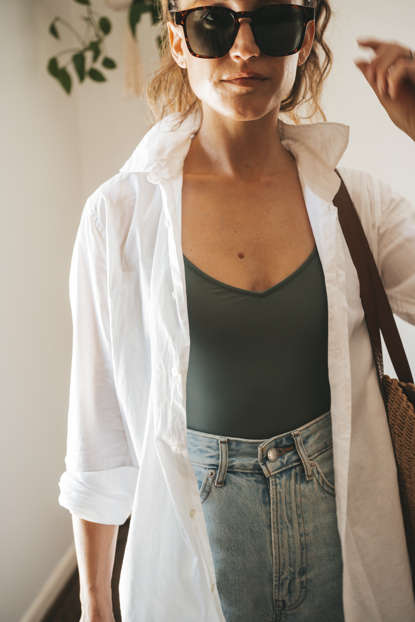 ashley wearing an olive green one piece swimsuit under a white button down, light wash denim shorts, and white sandals with tortoise shell sunglasses