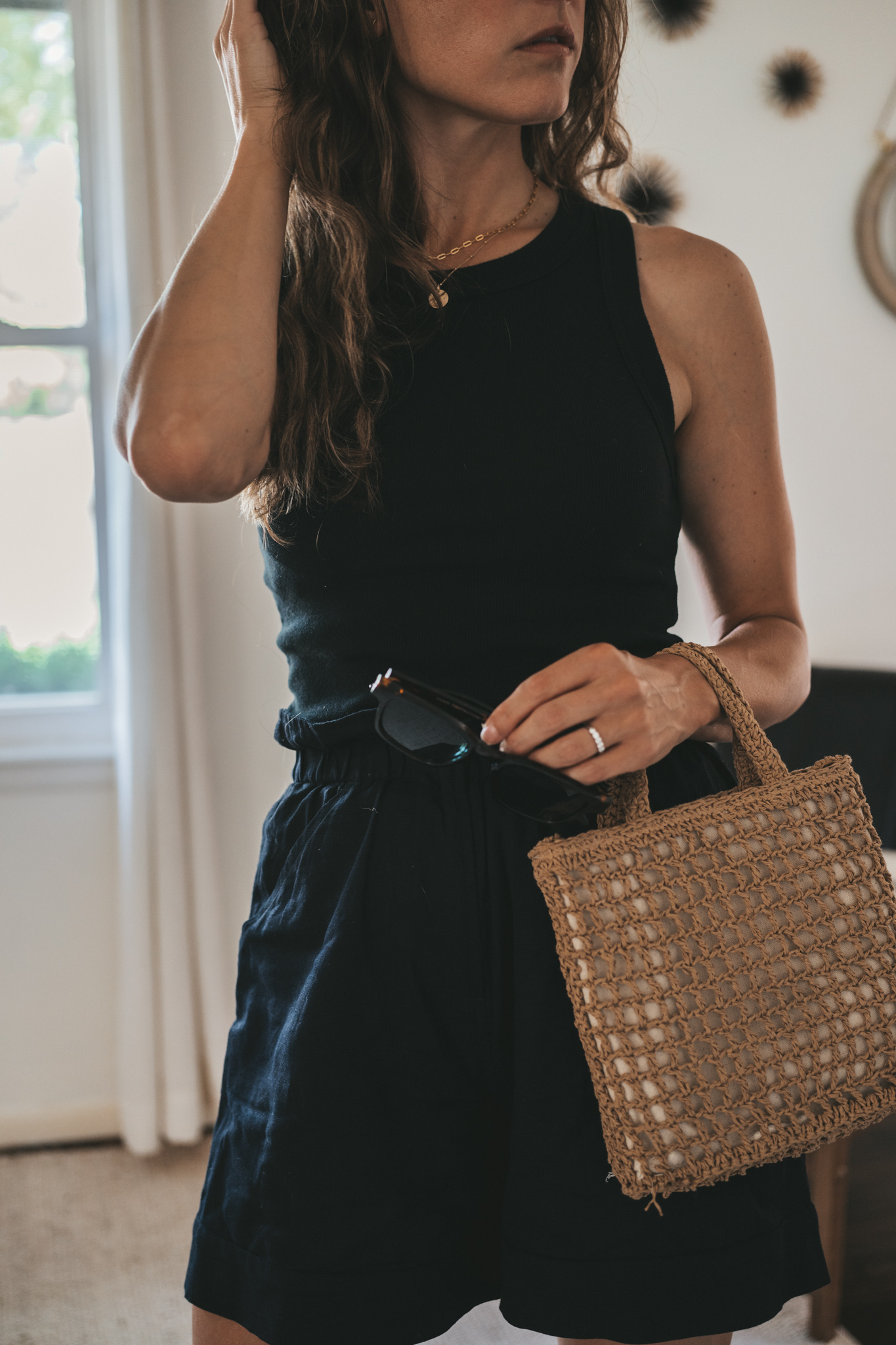 ashley wearing a black ribbed tank, black paperbag shorts, black strappy sandals, tortoise shell sunglasses, and a woven bag