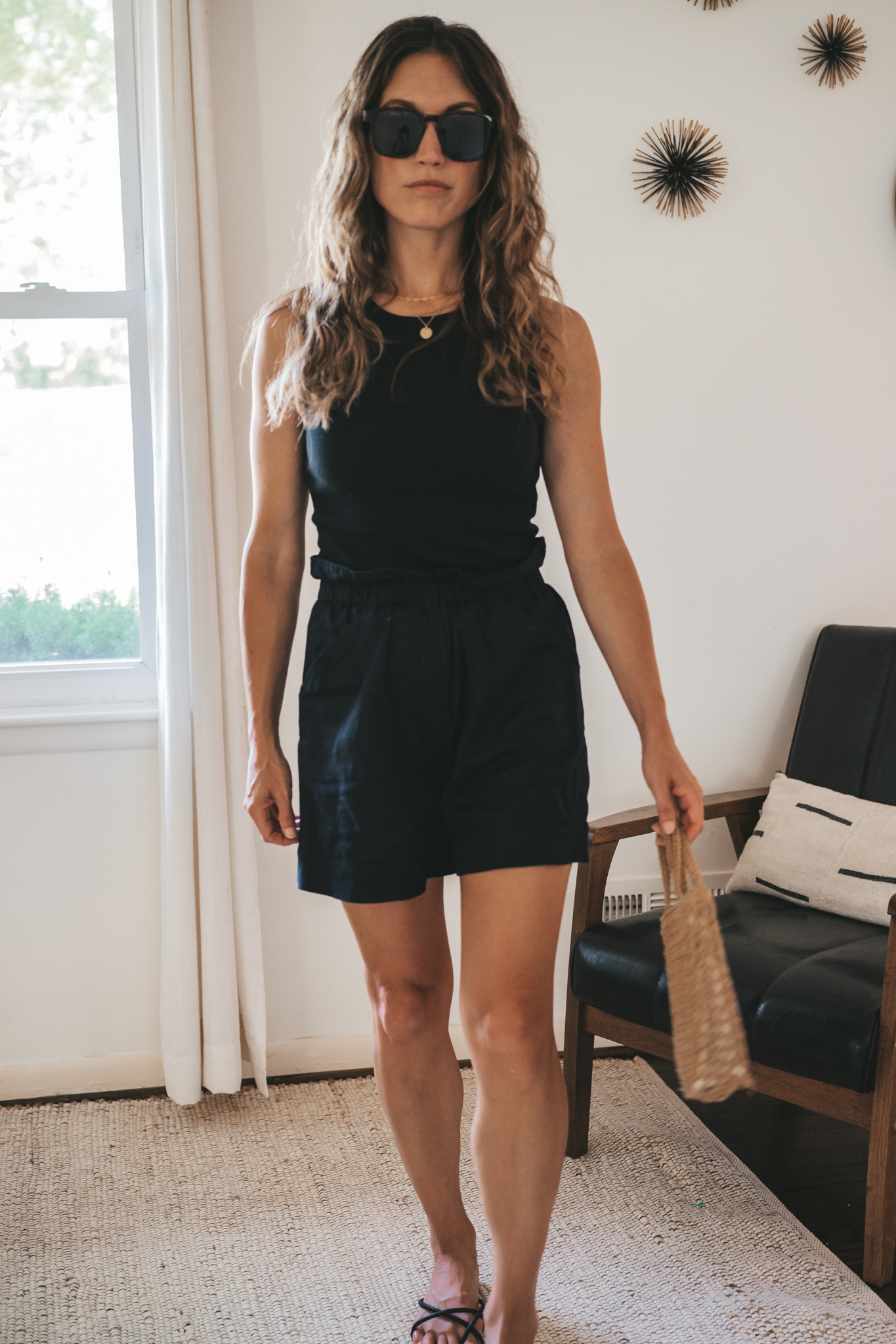 ashley wearing a black ribbed tank, black paperbag shorts, black strappy sandals, tortoise shell sunglasses, and a woven bag