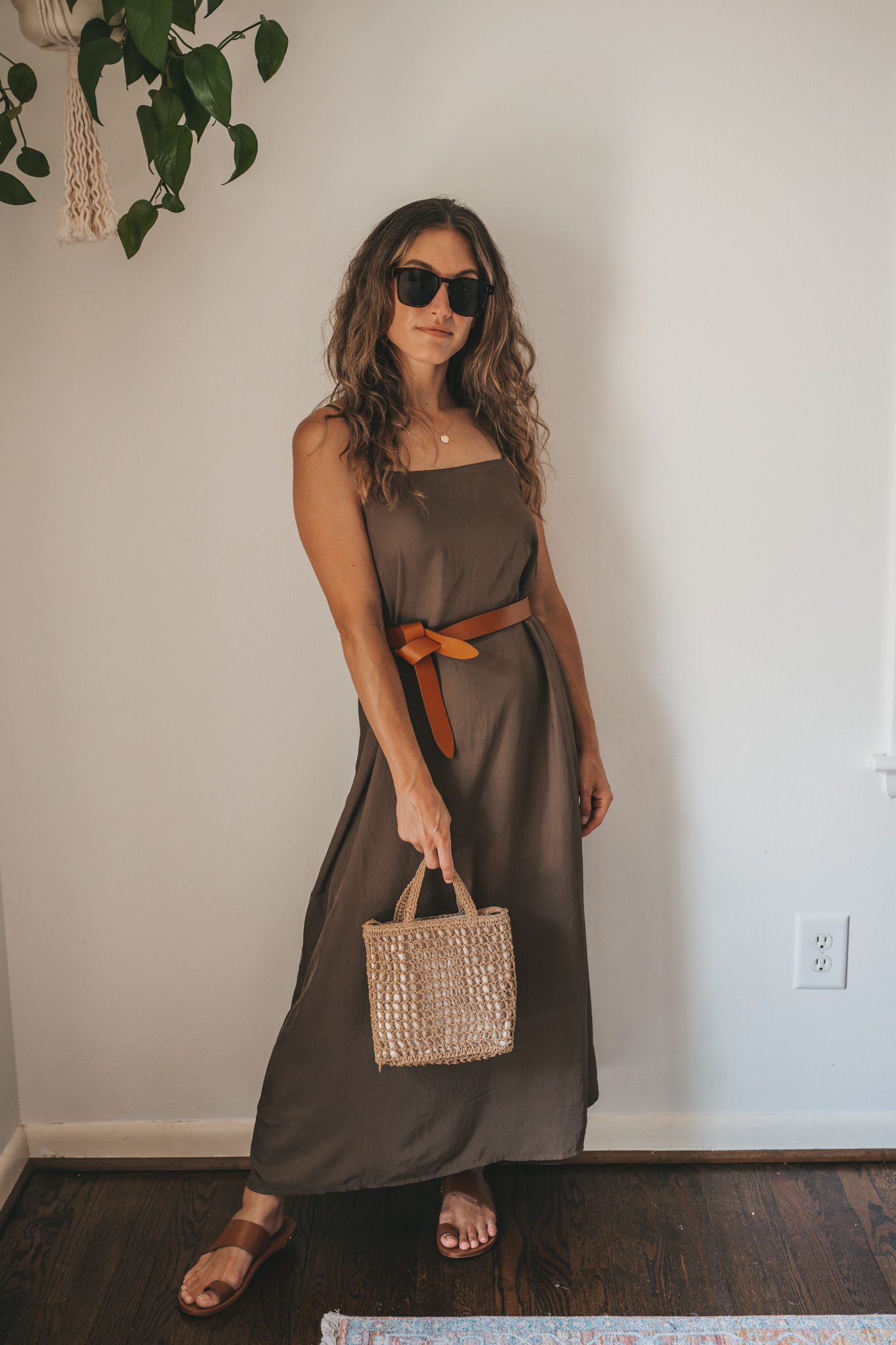 ashley wearing an olive green slip dress with a brown leather belt tied at the waist, brown leather sandals, a woven bag, and tortoise shell sunglasses
