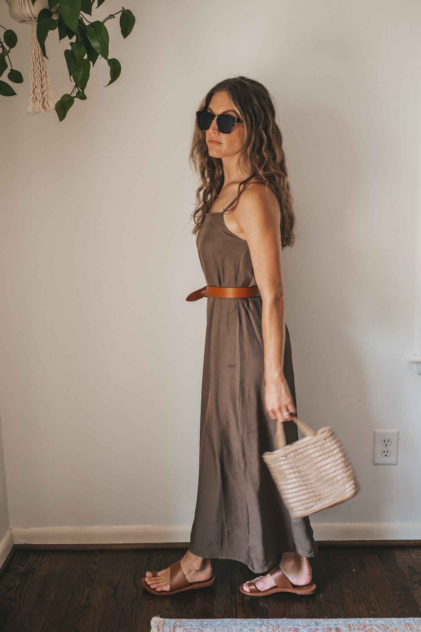 ashley wearing an olive green slip dress with a brown leather belt tied at the waist, brown leather sandals, a woven bag, and tortoise shell sunglasses