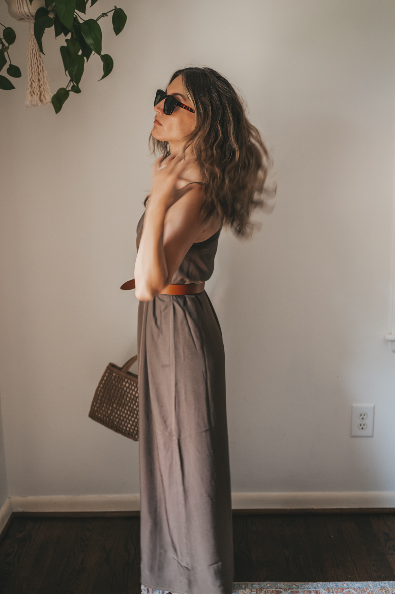 ashley wearing an olive green slip dress with a brown leather belt tied at the waist, brown leather sandals, a woven bag, and tortoise shell sunglasses