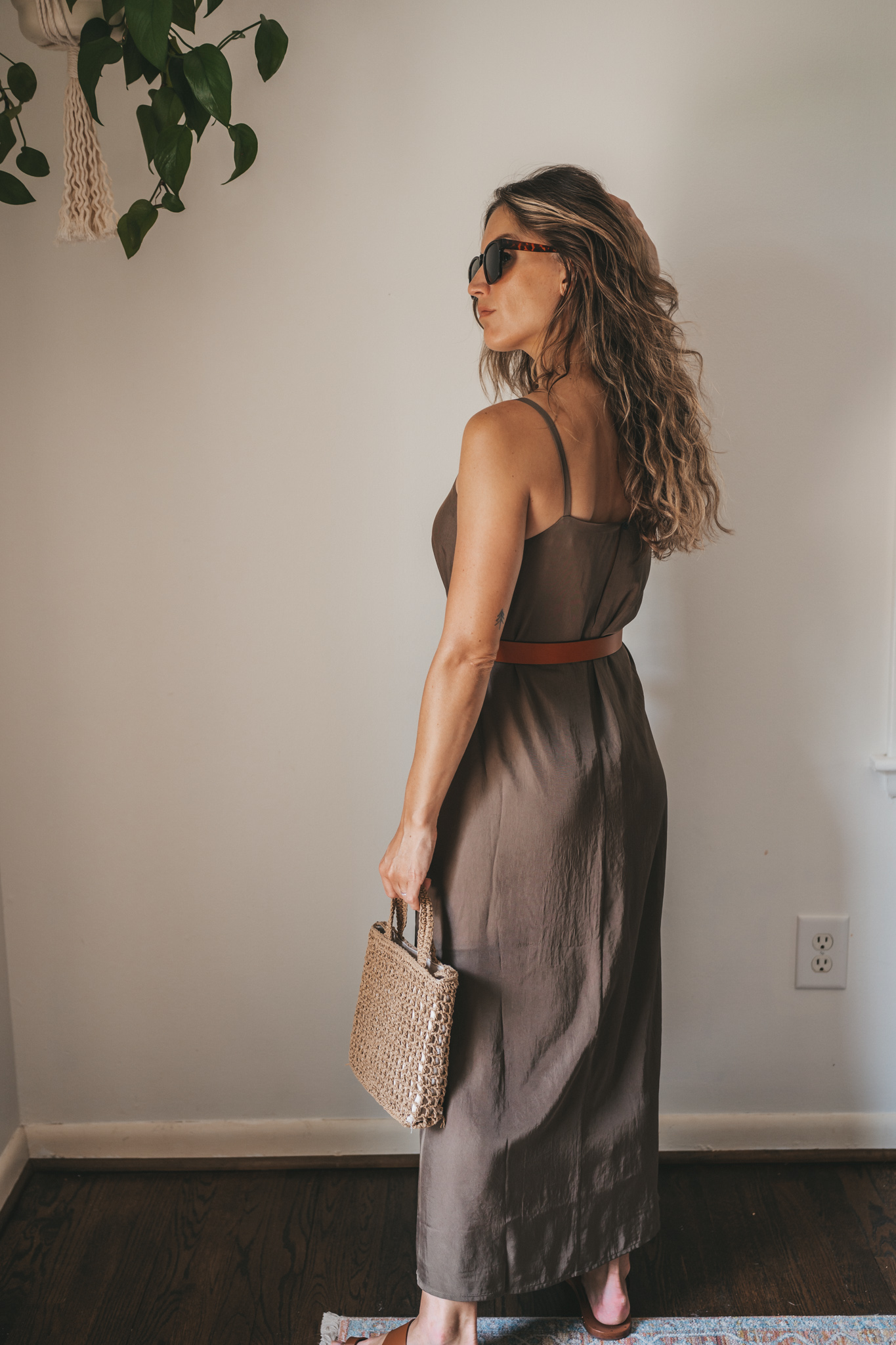 ashley wearing an olive green slip dress with a brown leather belt tied at the waist, brown leather sandals, a woven bag, and tortoise shell sunglasses