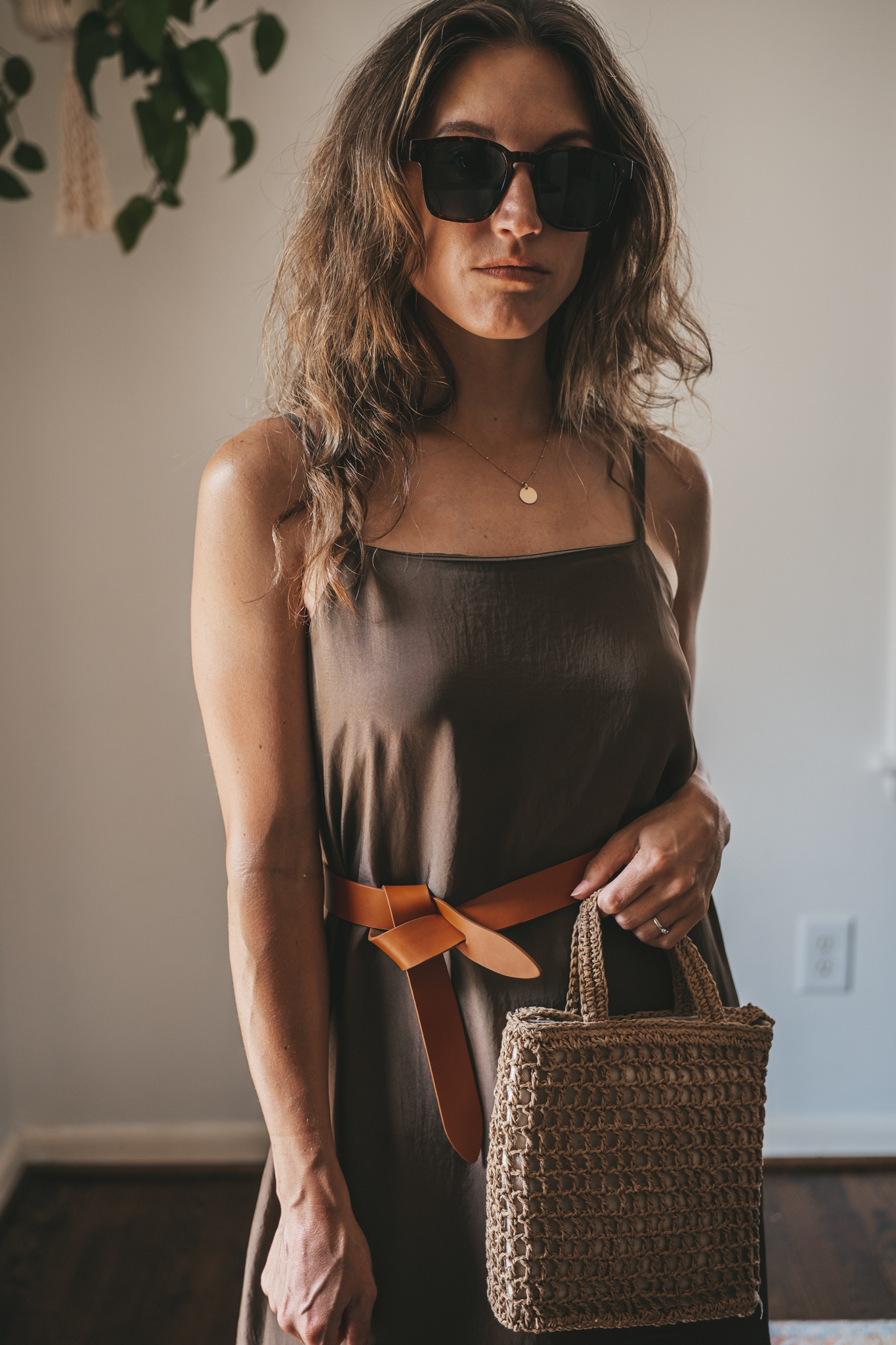 ashley wearing an olive green slip dress with a brown leather belt tied at the waist, brown leather sandals, a woven bag, and tortoise shell sunglasses