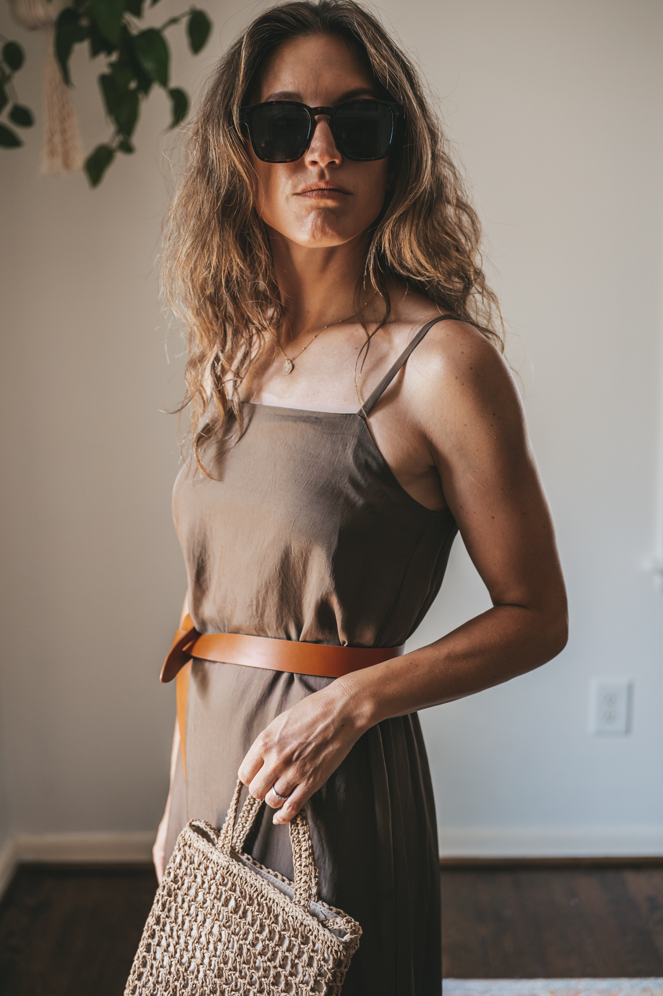 ashley wearing an olive green slip dress with a brown leather belt tied at the waist, brown leather sandals, a woven bag, and tortoise shell sunglasses