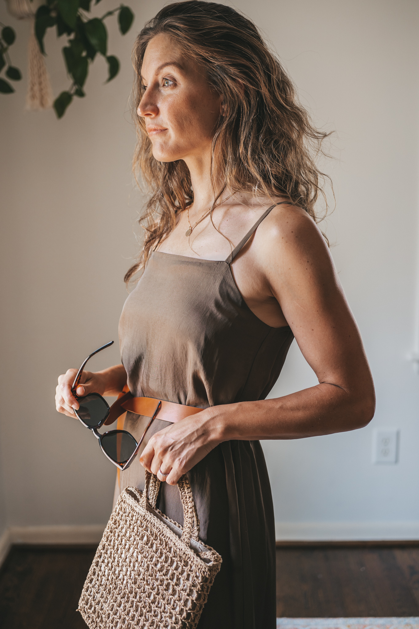 ashley wearing an olive green slip dress with a brown leather belt tied at the waist, brown leather sandals, a woven bag, and tortoise shell sunglasses