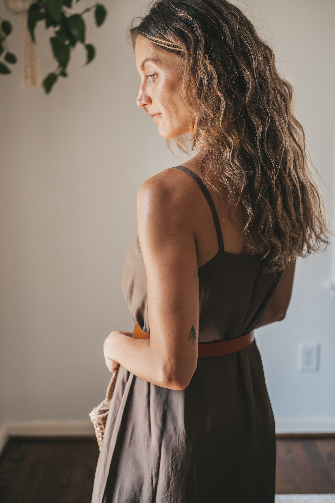 ashley wearing an olive green slip dress with a brown leather belt tied at the waist, brown leather sandals, a woven bag, and tortoise shell sunglasses
