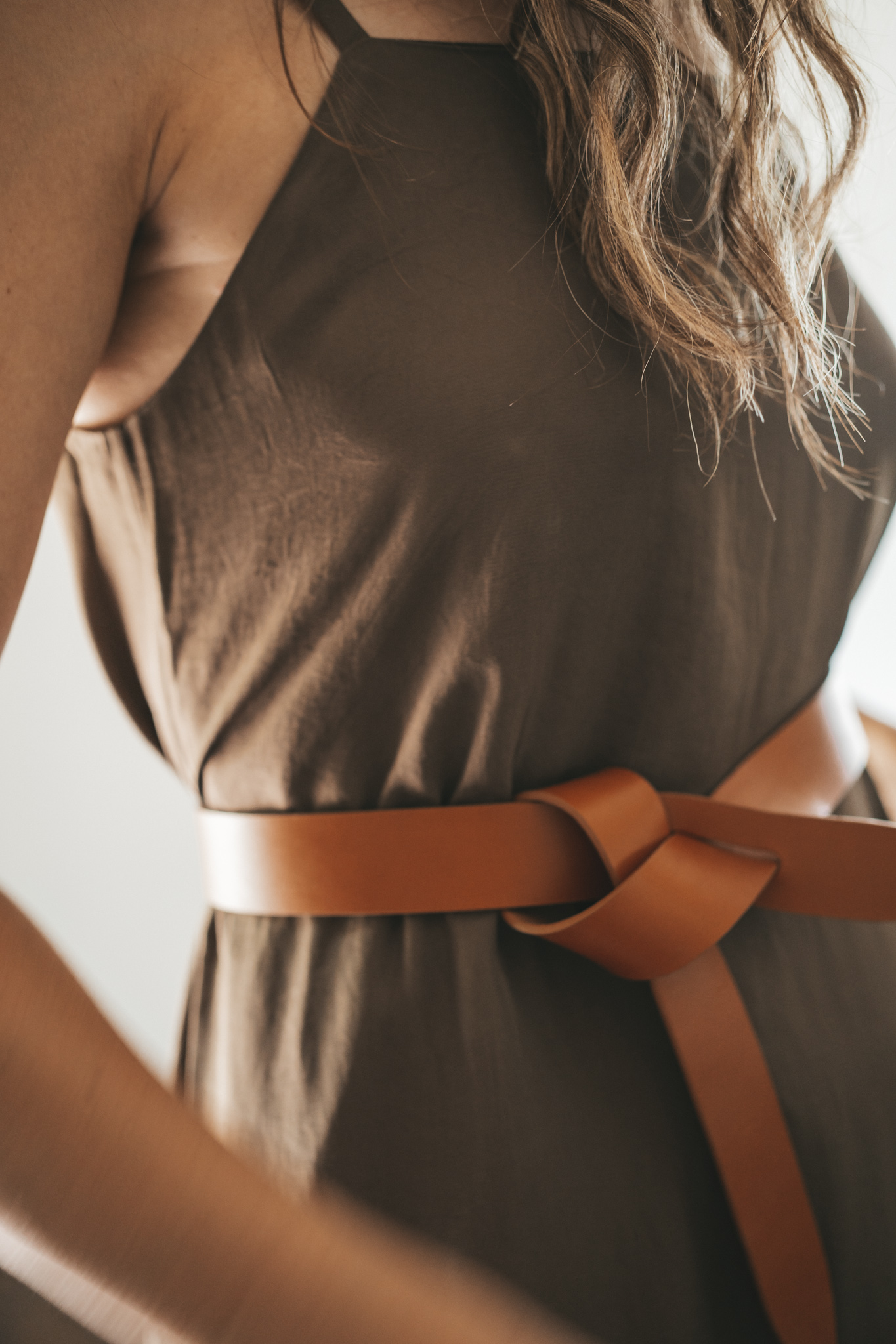 ashley wearing an olive green slip dress with a brown leather belt tied at the waist, brown leather sandals, a woven bag, and tortoise shell sunglasses