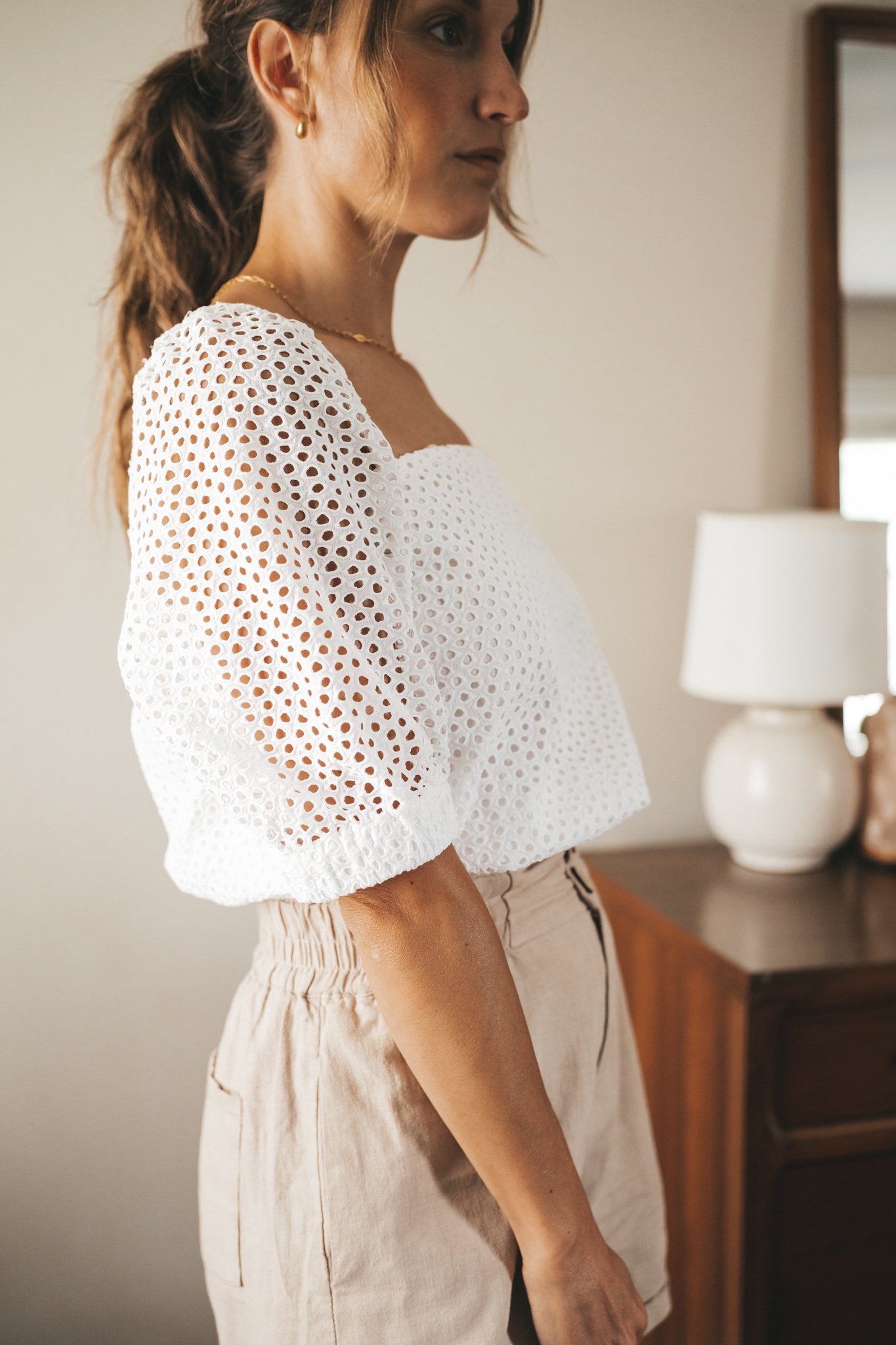 ashley wearing a white puff sleeve eyelet top, tan linen blend shorts, nude sandals, and a gray shoulder bag