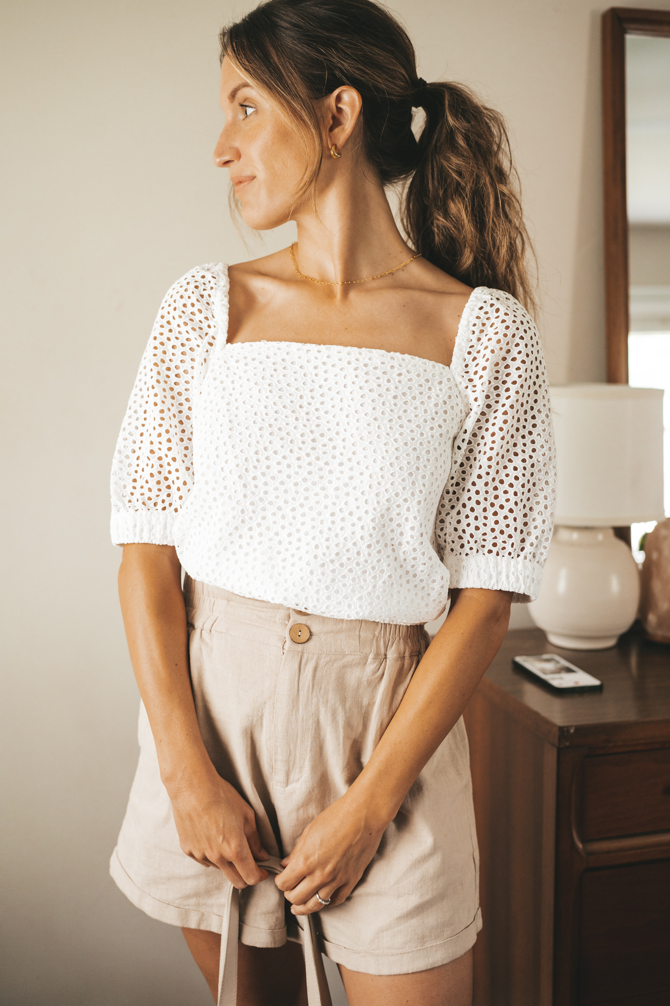ashley wearing a white puff sleeve eyelet top, tan linen blend shorts, nude sandals, and a gray shoulder bag
