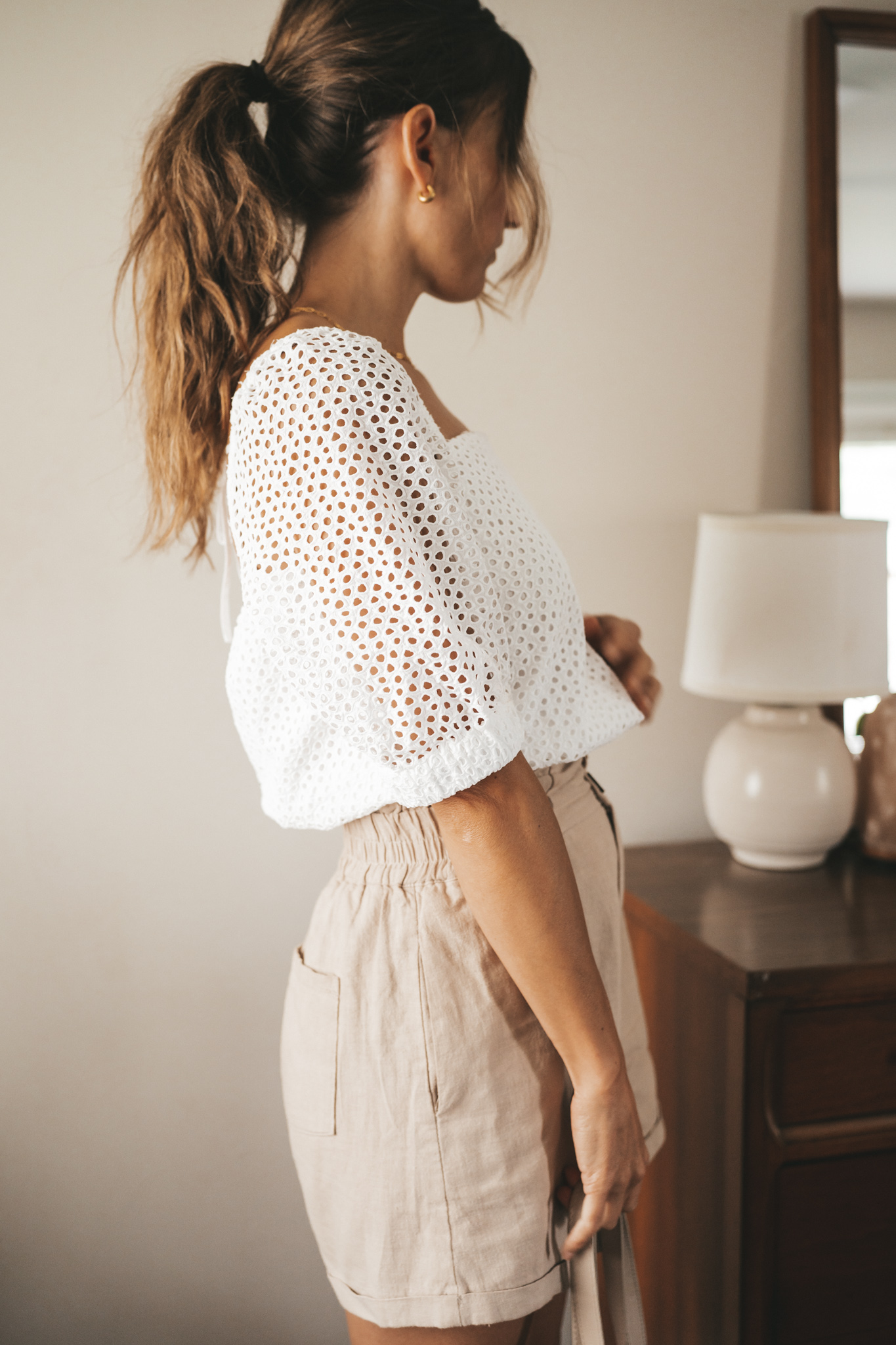 ashley wearing a white puff sleeve eyelet top, tan linen blend shorts, nude sandals, and a gray shoulder bag