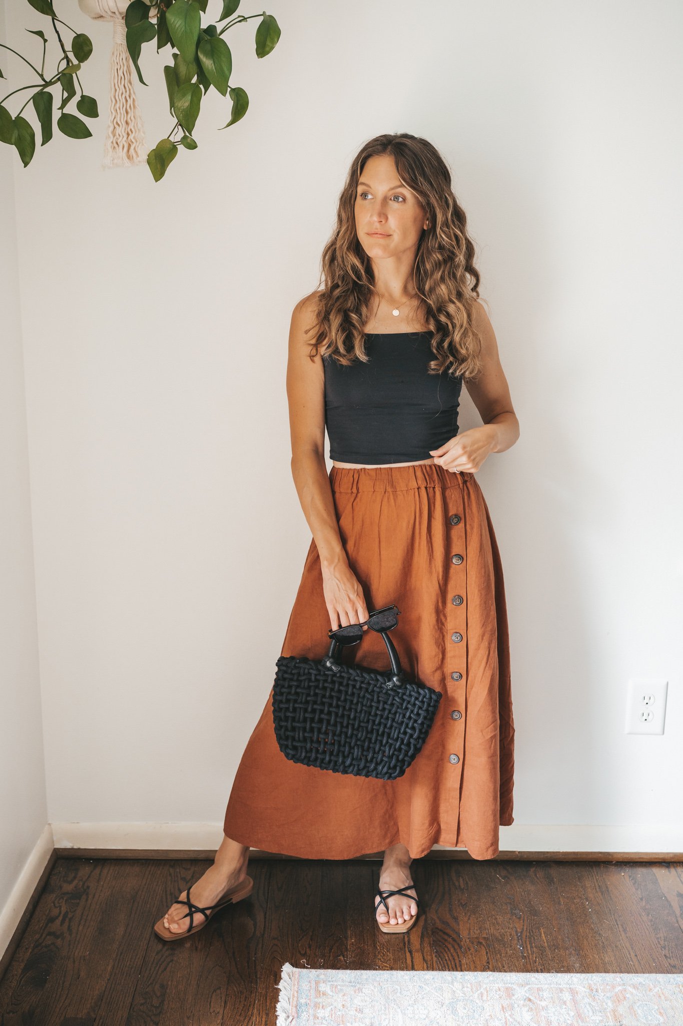 ashley wearing a cropped black tank, dark orange midi skirt with buttons down the front, black strappy sandals, a gold pendant necklace, and a black woven bag