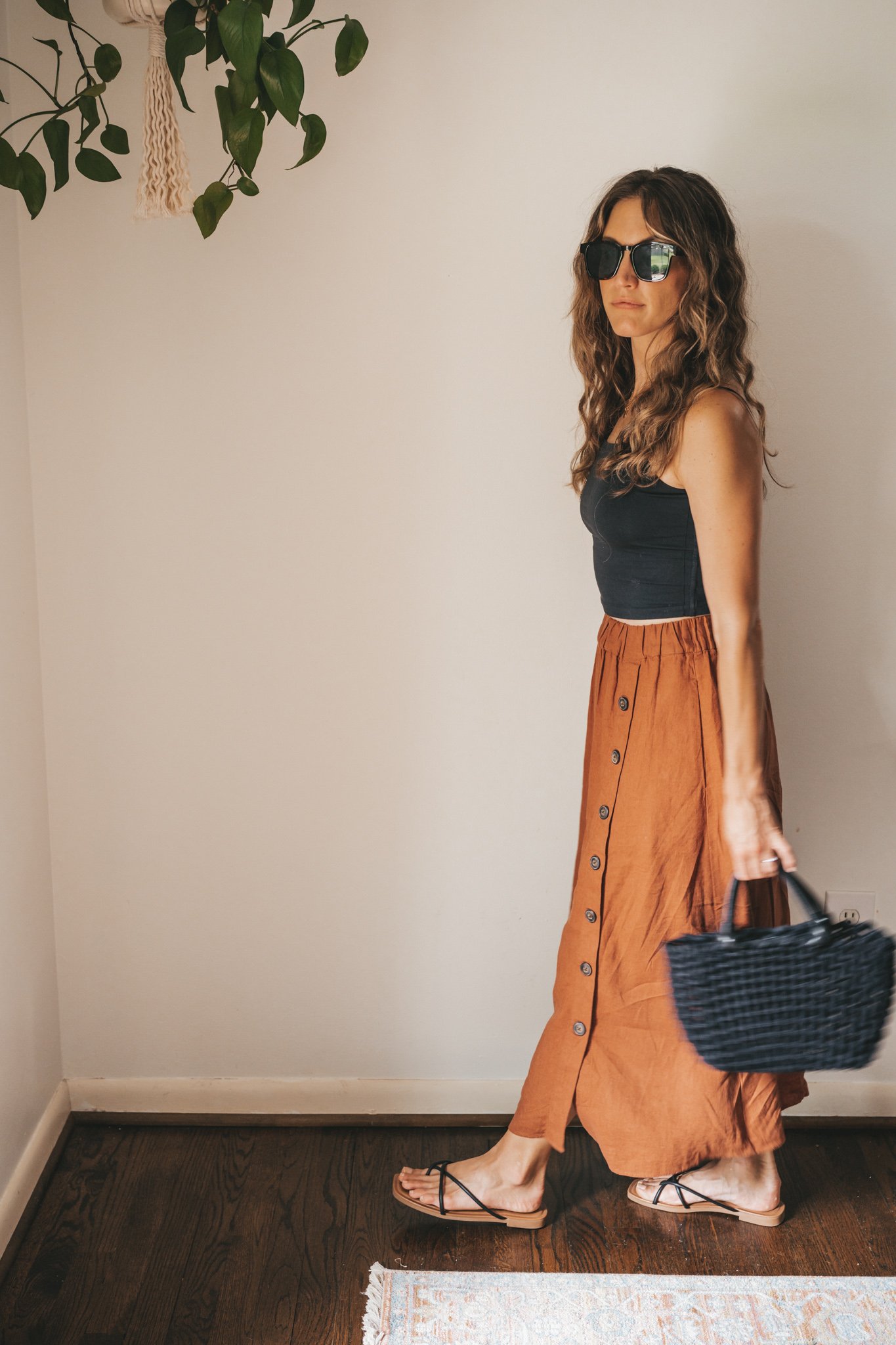 ashley wearing a cropped black tank, dark orange midi skirt with buttons down the front, black strappy sandals, a gold pendant necklace, and a black woven bag