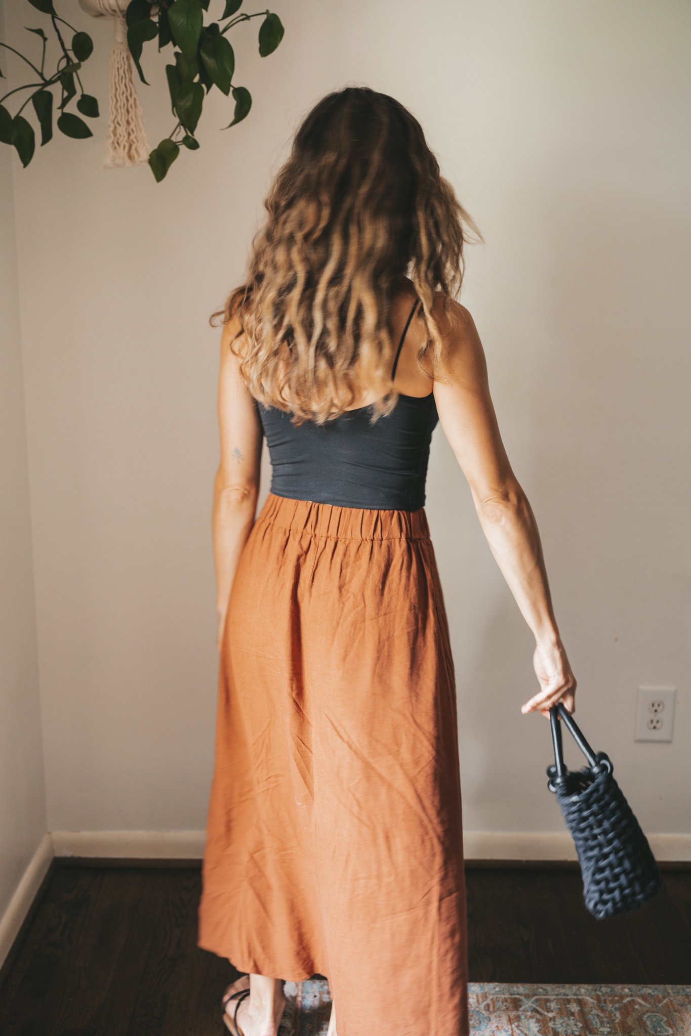 ashley wearing a cropped black tank, dark orange midi skirt with buttons down the front, black strappy sandals, a gold pendant necklace, and a black woven bag