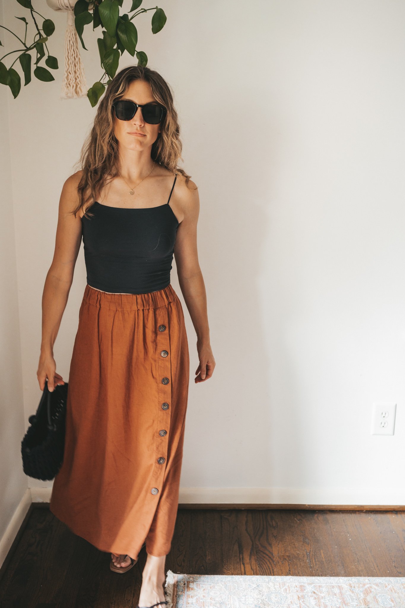 ashley wearing a cropped black tank, dark orange midi skirt with buttons down the front, black strappy sandals, a gold pendant necklace, and a black woven bag
