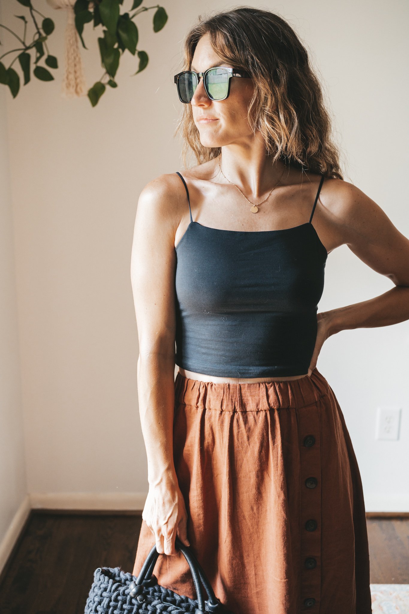 ashley wearing a cropped black tank, dark orange midi skirt with buttons down the front, black strappy sandals, a gold pendant necklace, and a black woven bag