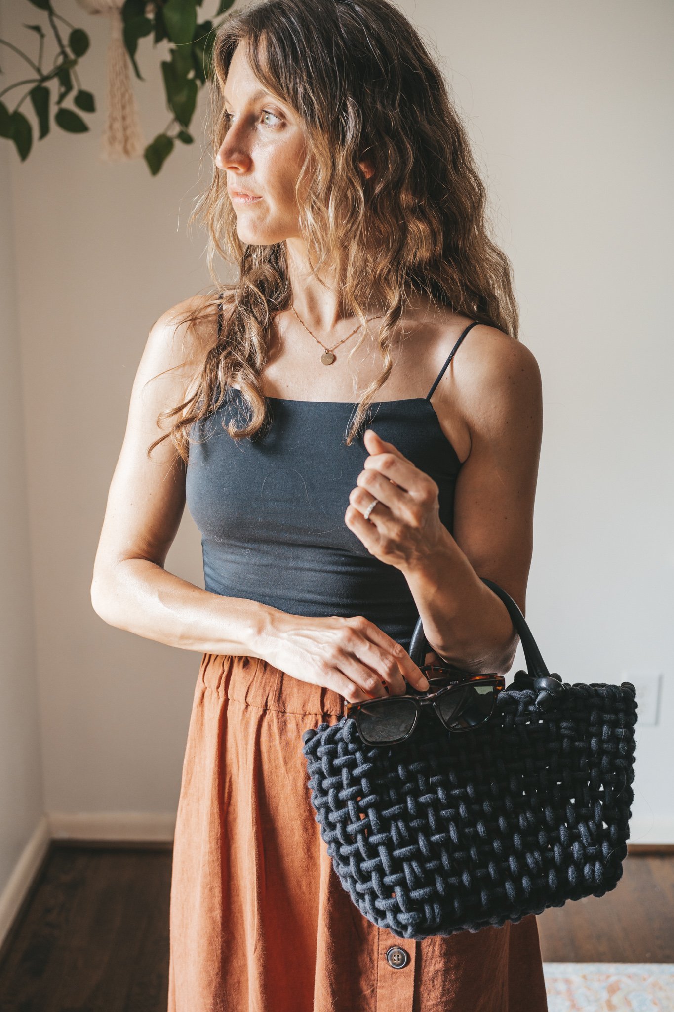 ashley wearing a cropped black tank, dark orange midi skirt with buttons down the front, black strappy sandals, a gold pendant necklace, and a black woven bag