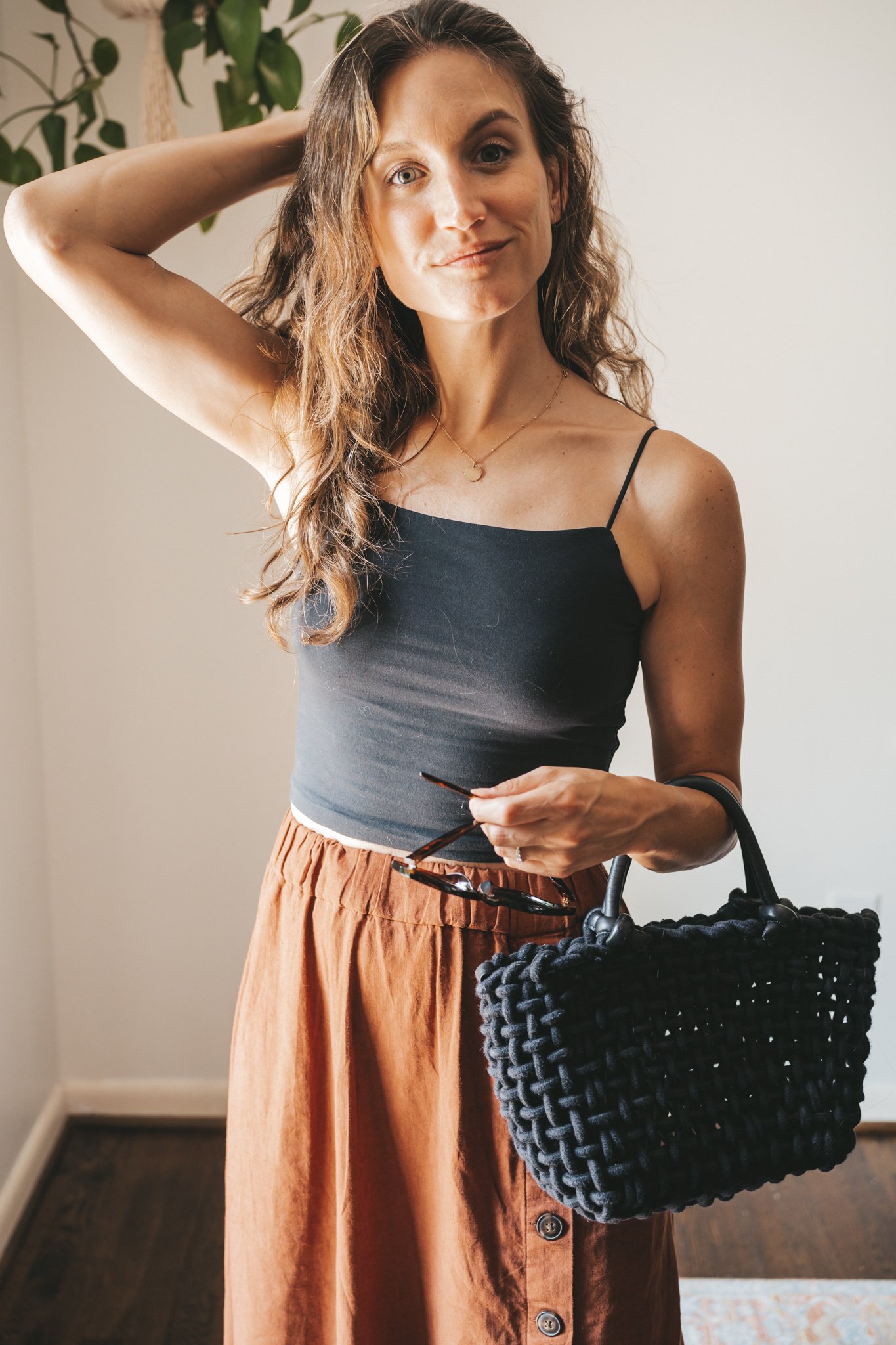 ashley wearing a cropped black tank, dark orange midi skirt with buttons down the front, black strappy sandals, a gold pendant necklace, and a black woven bag