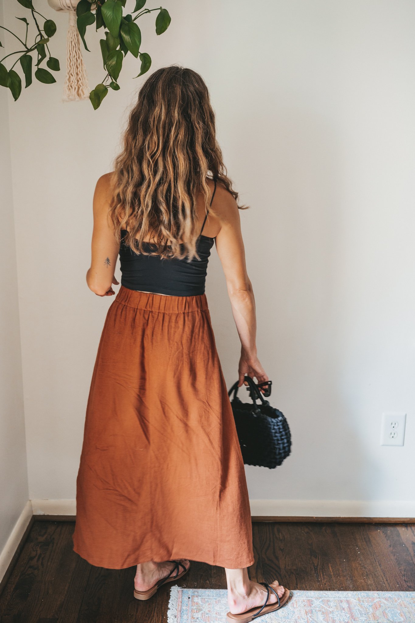 ashley wearing a cropped black tank, dark orange midi skirt with buttons down the front, black strappy sandals, a gold pendant necklace, and a black woven bag