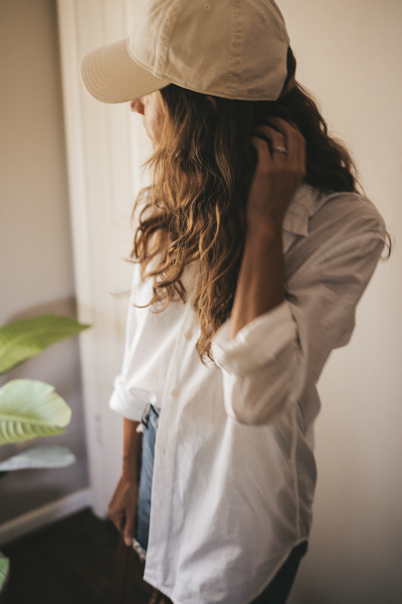 ashley wearing a white button down, denim shorts, white sandals, a woven tote bag, and a cream colored baseball cap.