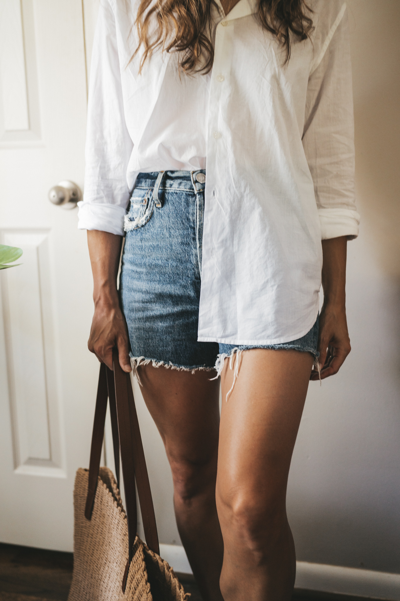 ashley wearing a white button down, denim shorts, white sandals, a woven tote bag, and a cream colored baseball cap.