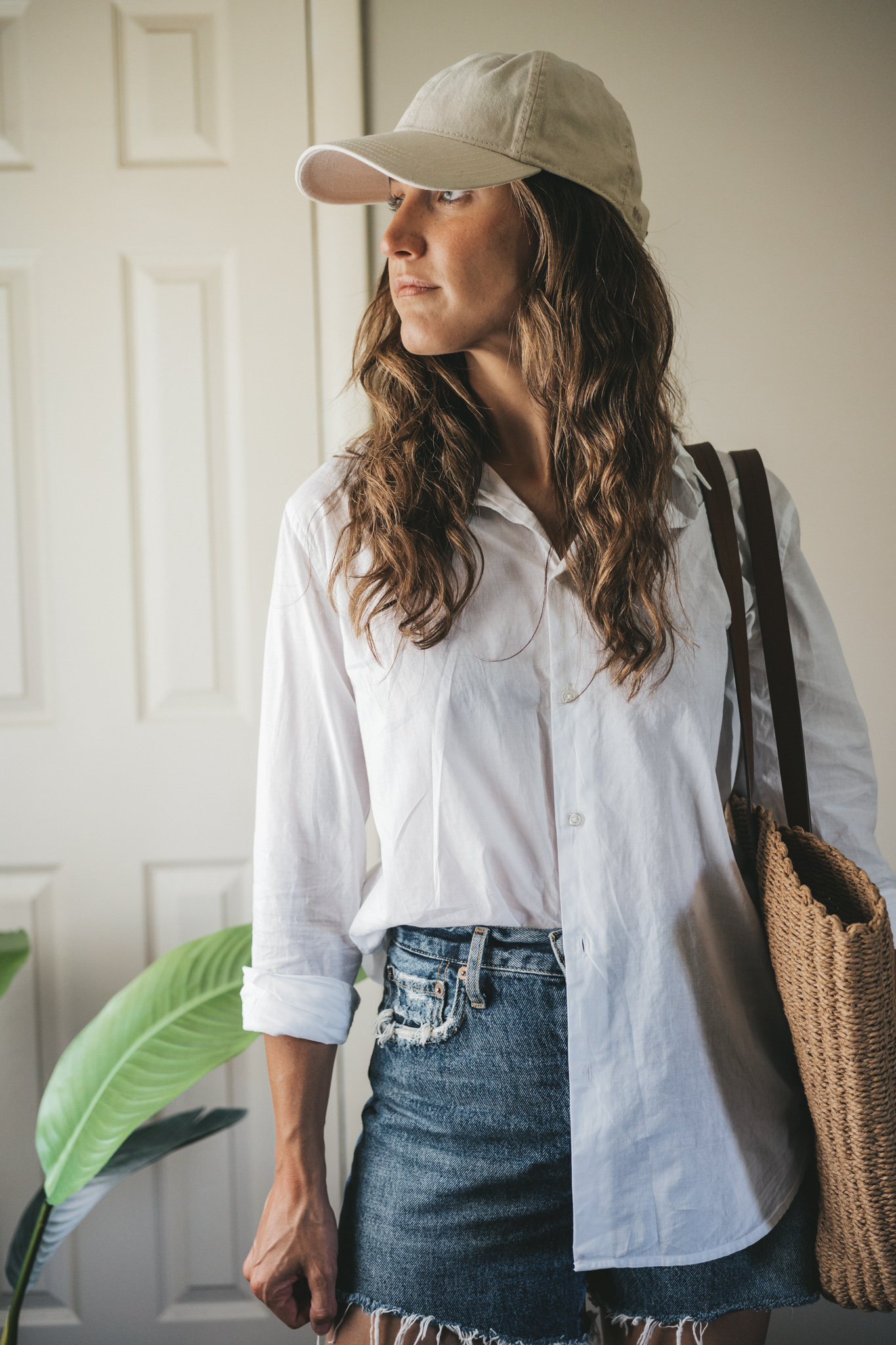 ashley wearing a white button down, denim shorts, white sandals, a woven tote bag, and a cream colored baseball cap.