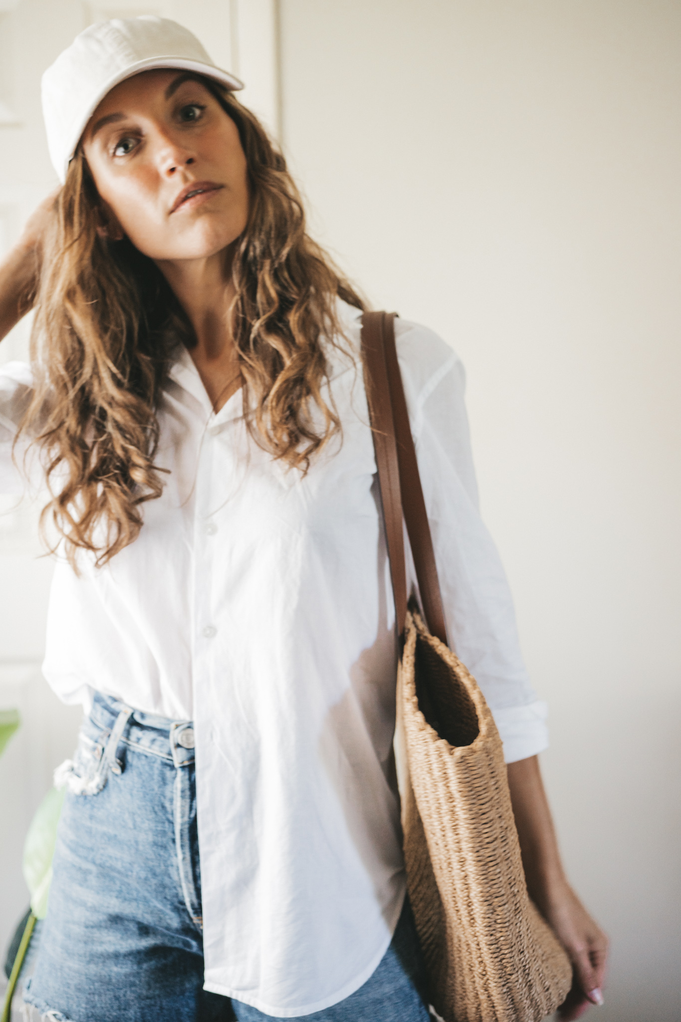 ashley wearing a white button down, denim shorts, white sandals, a woven tote bag, and a cream colored baseball cap.
