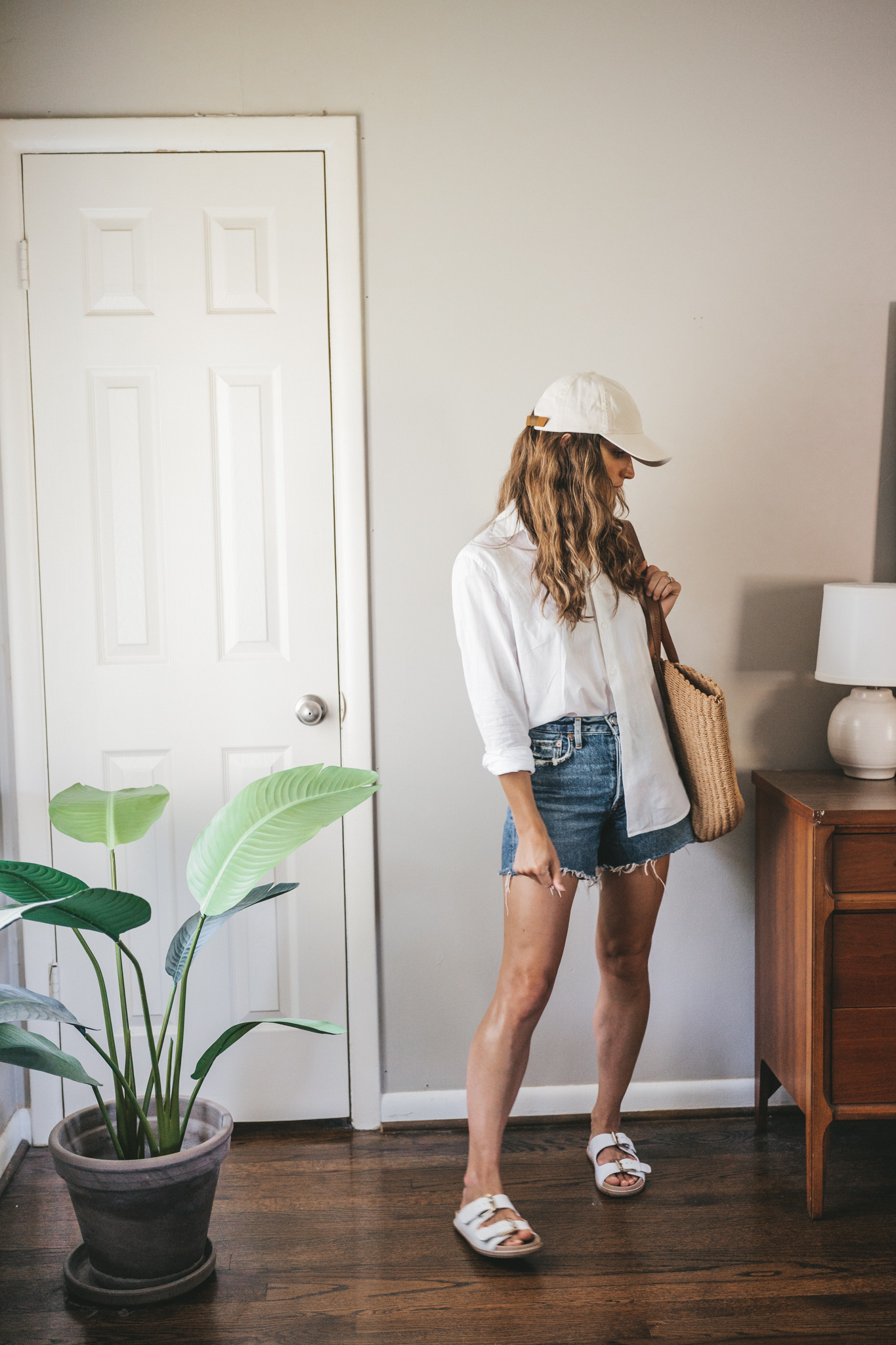 ashley wearing a white button down, denim shorts, white sandals, a woven tote bag, and a cream colored baseball cap.
