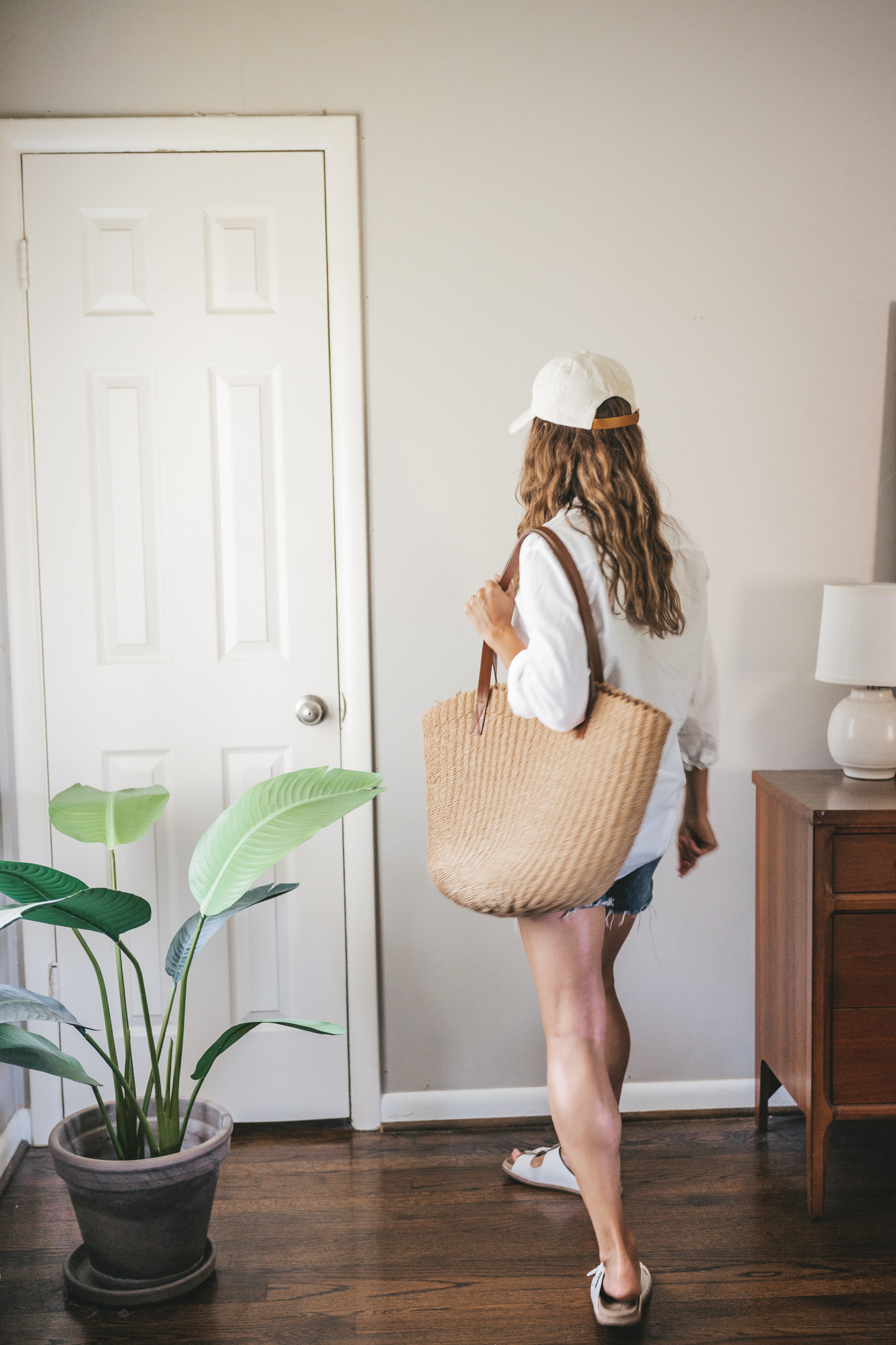 ashley wearing a white button down, denim shorts, white sandals, a woven tote bag, and a cream colored baseball cap.