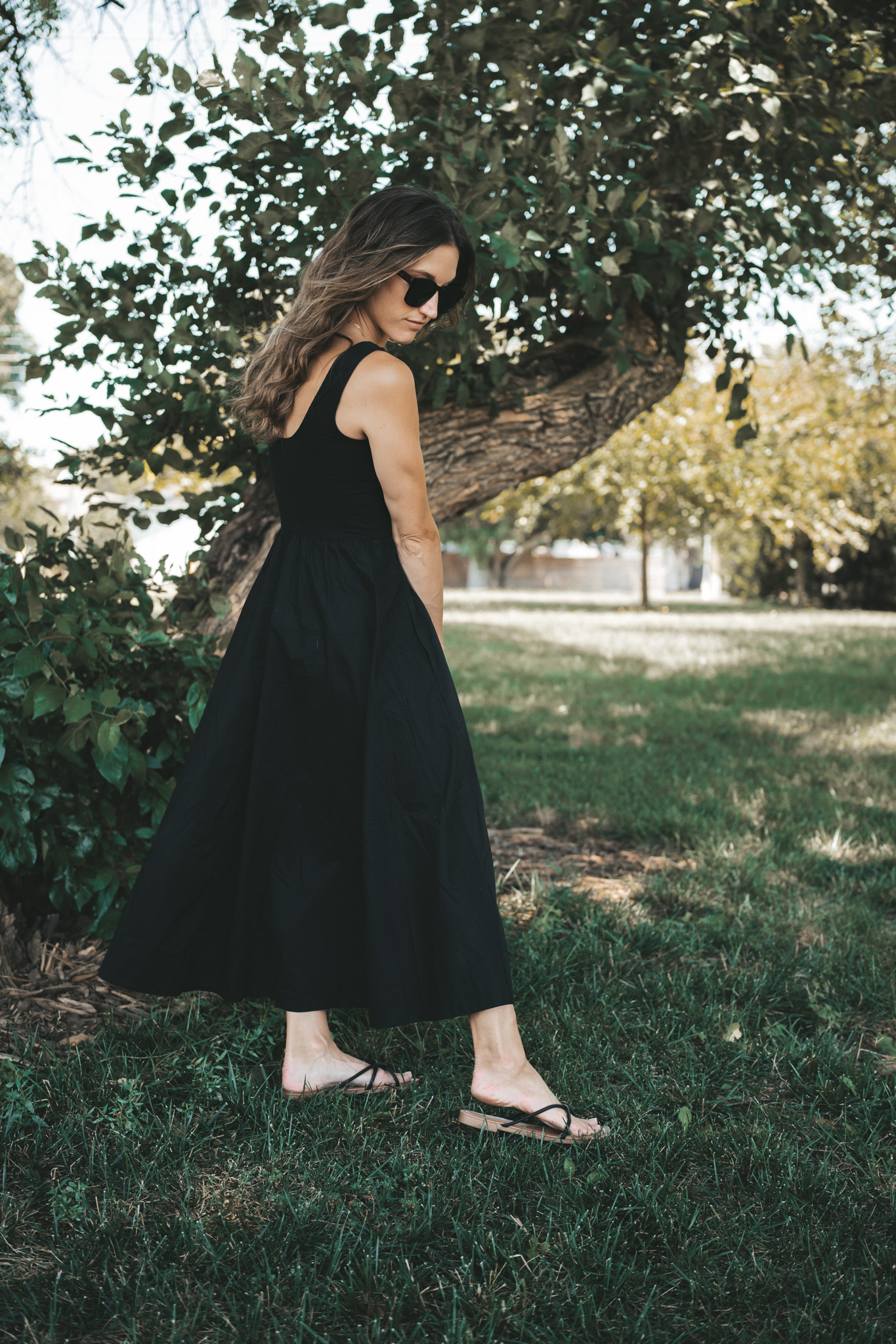 ashley wearing a black a-line midi dress, black strappy sandals, tortoise shell sunglasses, and a woven bag standing outside next to a tree