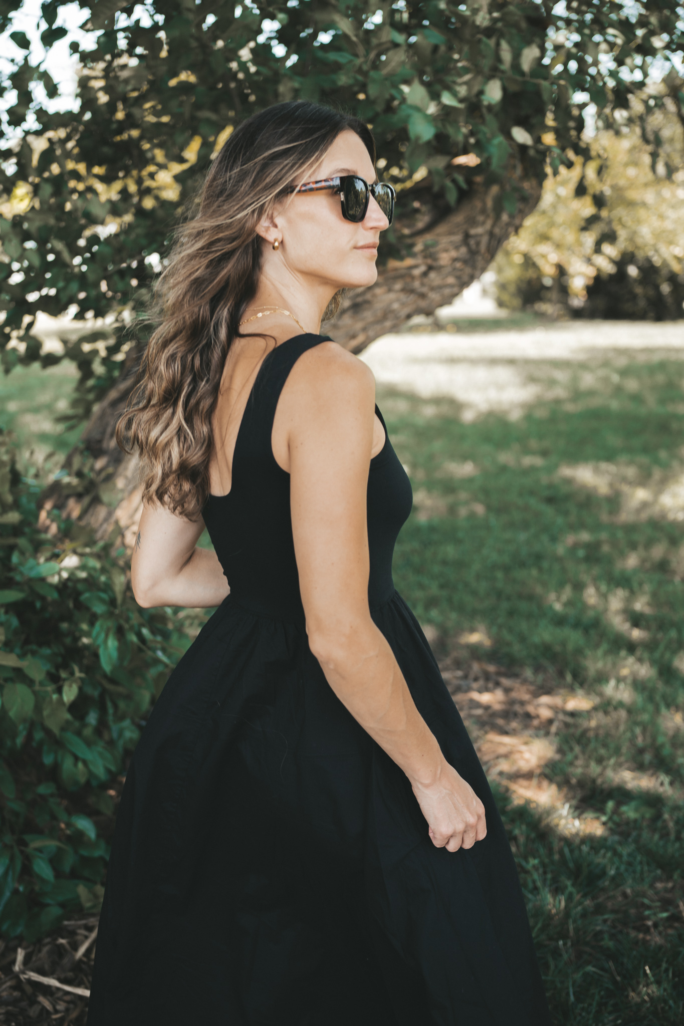 ashley wearing a black a-line midi dress, black strappy sandals, tortoise shell sunglasses, and a woven bag standing outside next to a tree