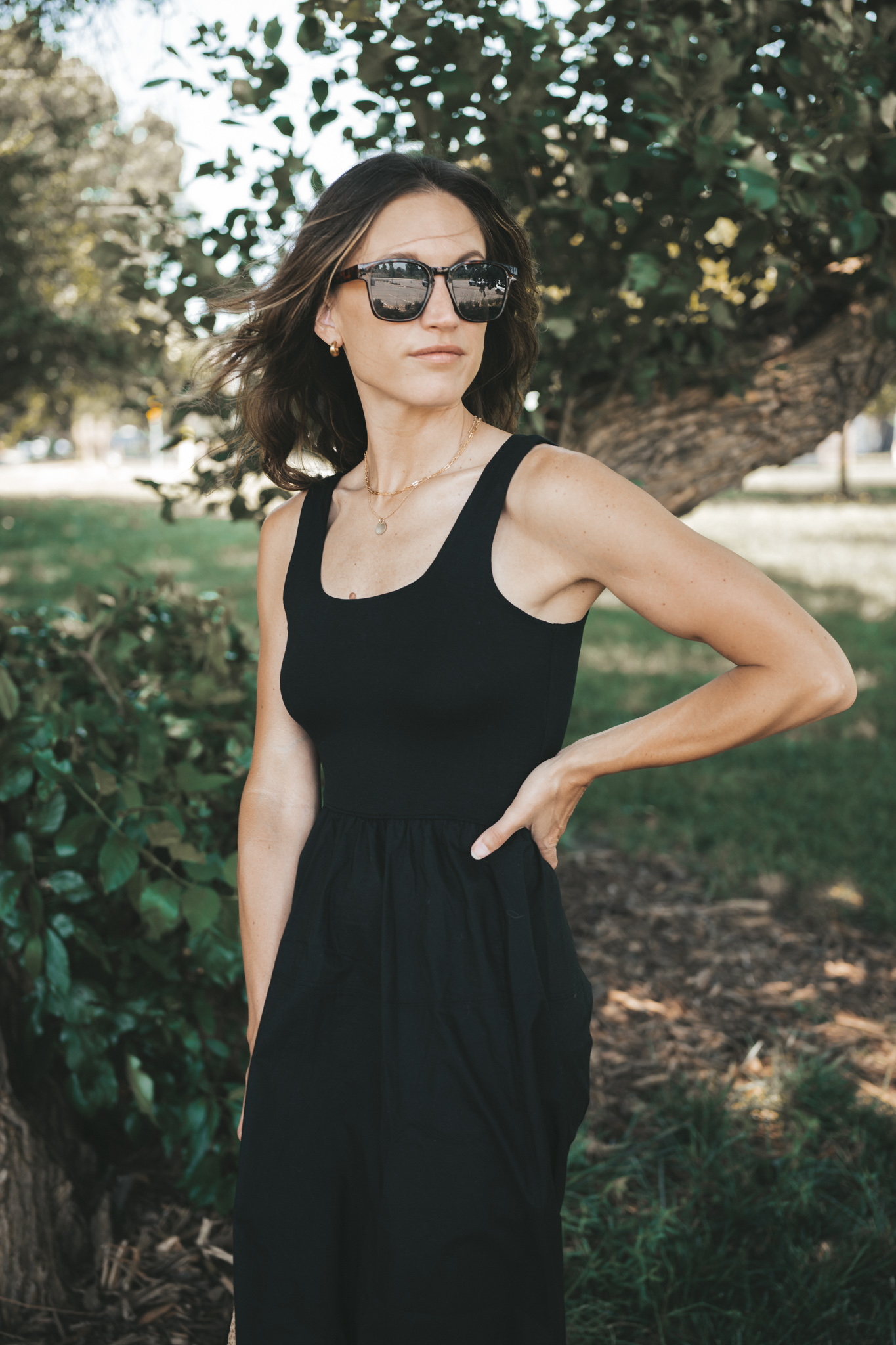 ashley wearing a black a-line midi dress, black strappy sandals, tortoise shell sunglasses, and a woven bag standing outside next to a tree