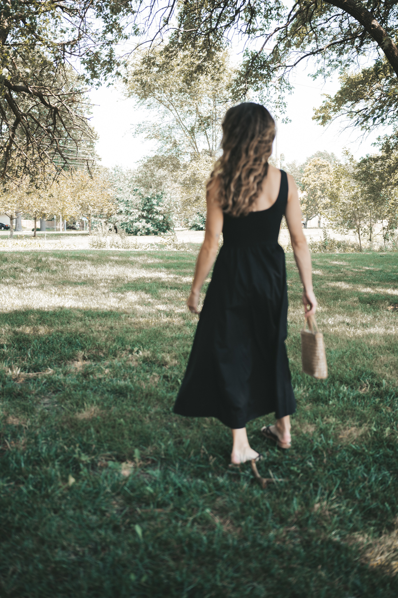 ashley wearing a black a-line midi dress, black strappy sandals, tortoise shell sunglasses, and a woven bag standing outside next to a tree