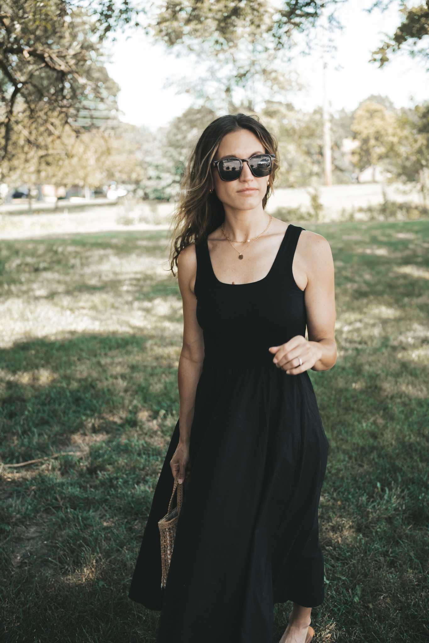 ashley wearing a black a-line midi dress, black strappy sandals, tortoise shell sunglasses, and a woven bag standing outside next to a tree