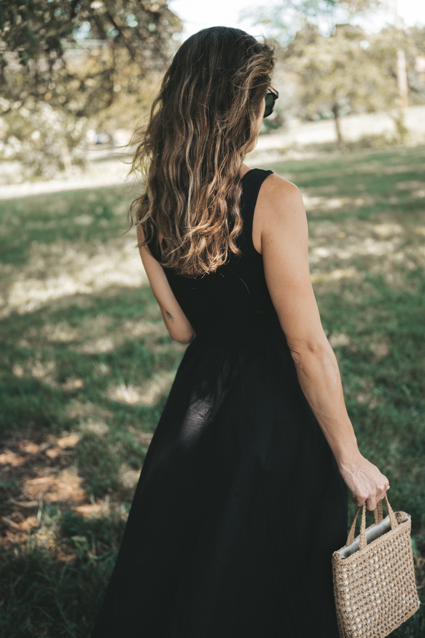 ashley wearing a black a-line midi dress, black strappy sandals, tortoise shell sunglasses, and a woven bag standing outside next to a tree