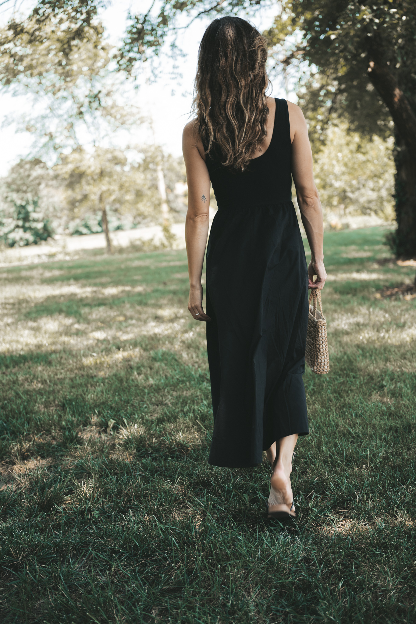 ashley wearing a black a-line midi dress, black strappy sandals, tortoise shell sunglasses, and a woven bag standing outside next to a tree
