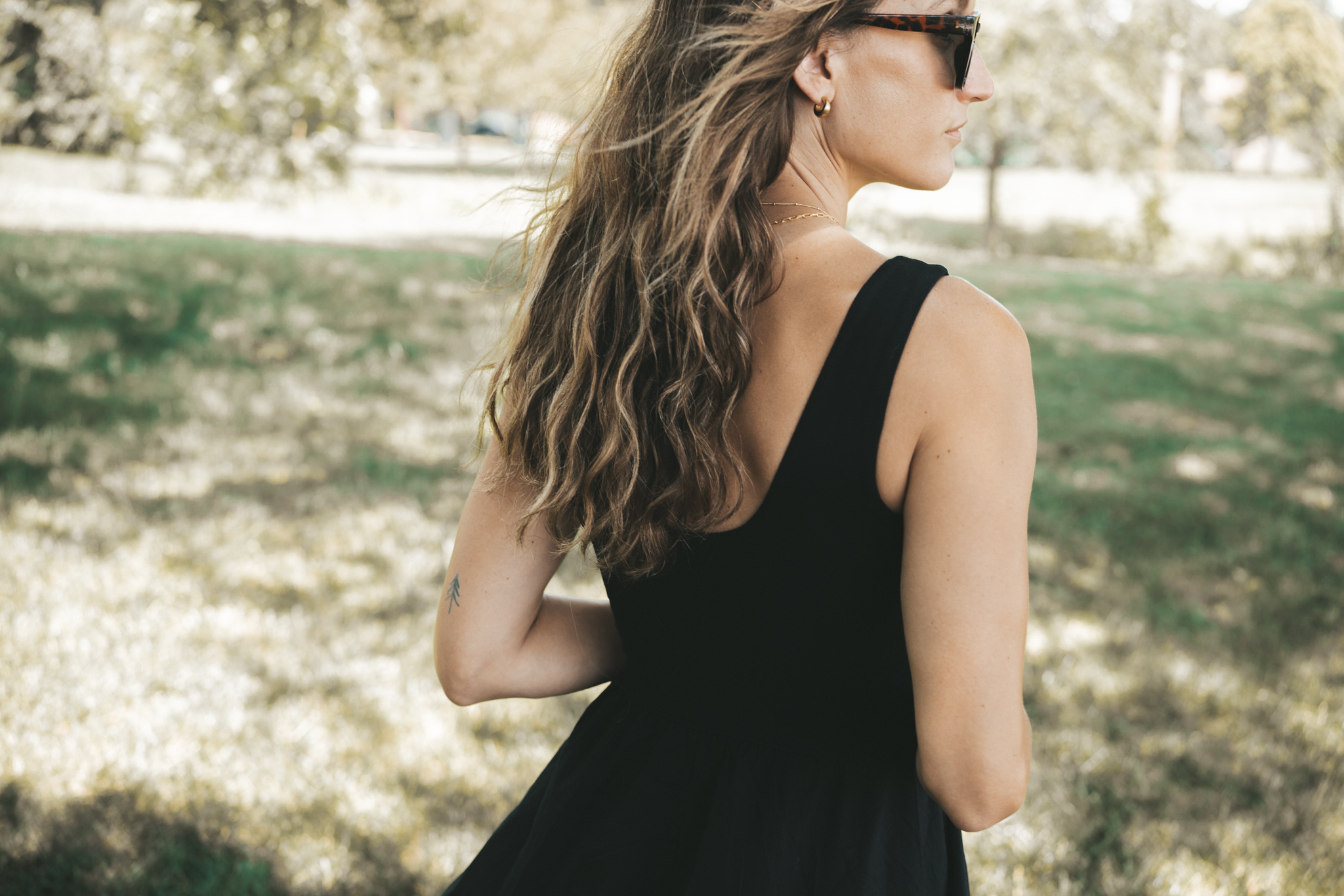 ashley wearing a black a-line midi dress, black strappy sandals, tortoise shell sunglasses, and a woven bag standing outside next to a tree